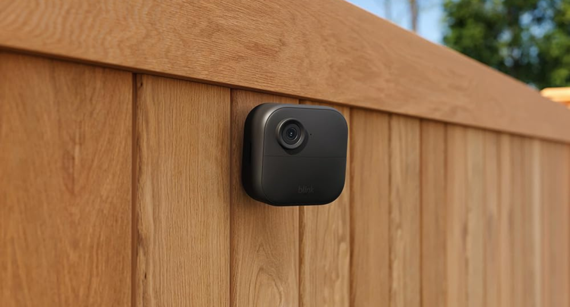 A black Blink Outdoor 4 camera mounted on a wooden fence.