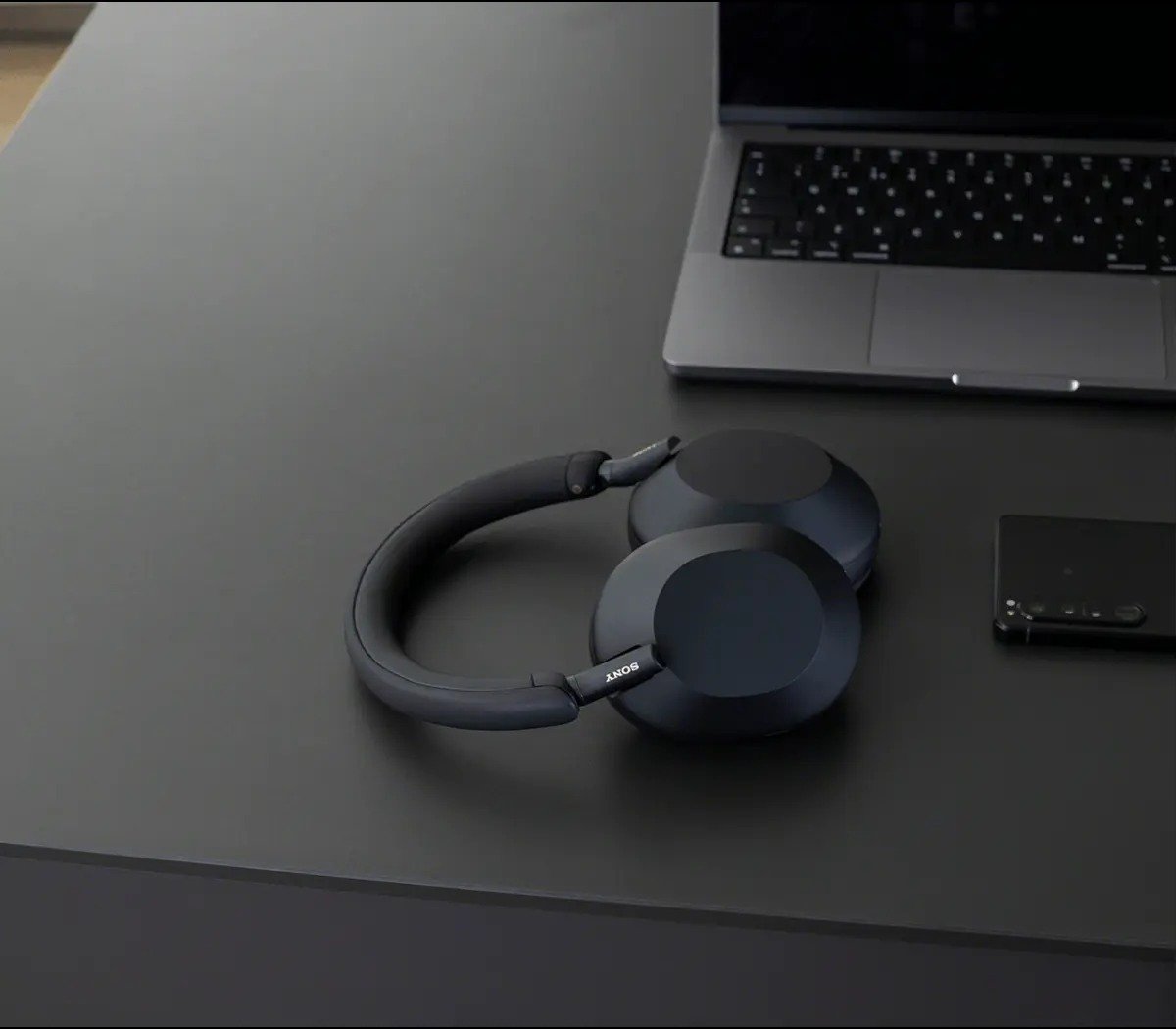 a pair of sony headphones sits on a black desk with a laptop and cell phone