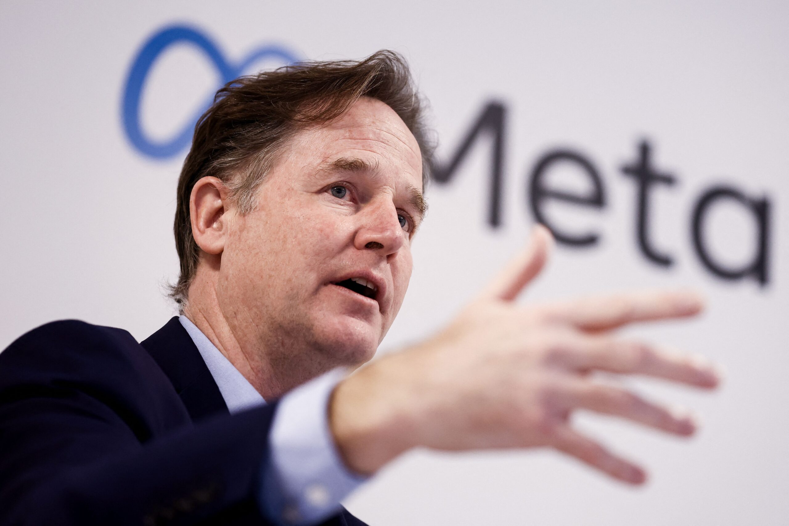 Nick Clegg gestures onstage in front of a backdrop with the Meta logo. 