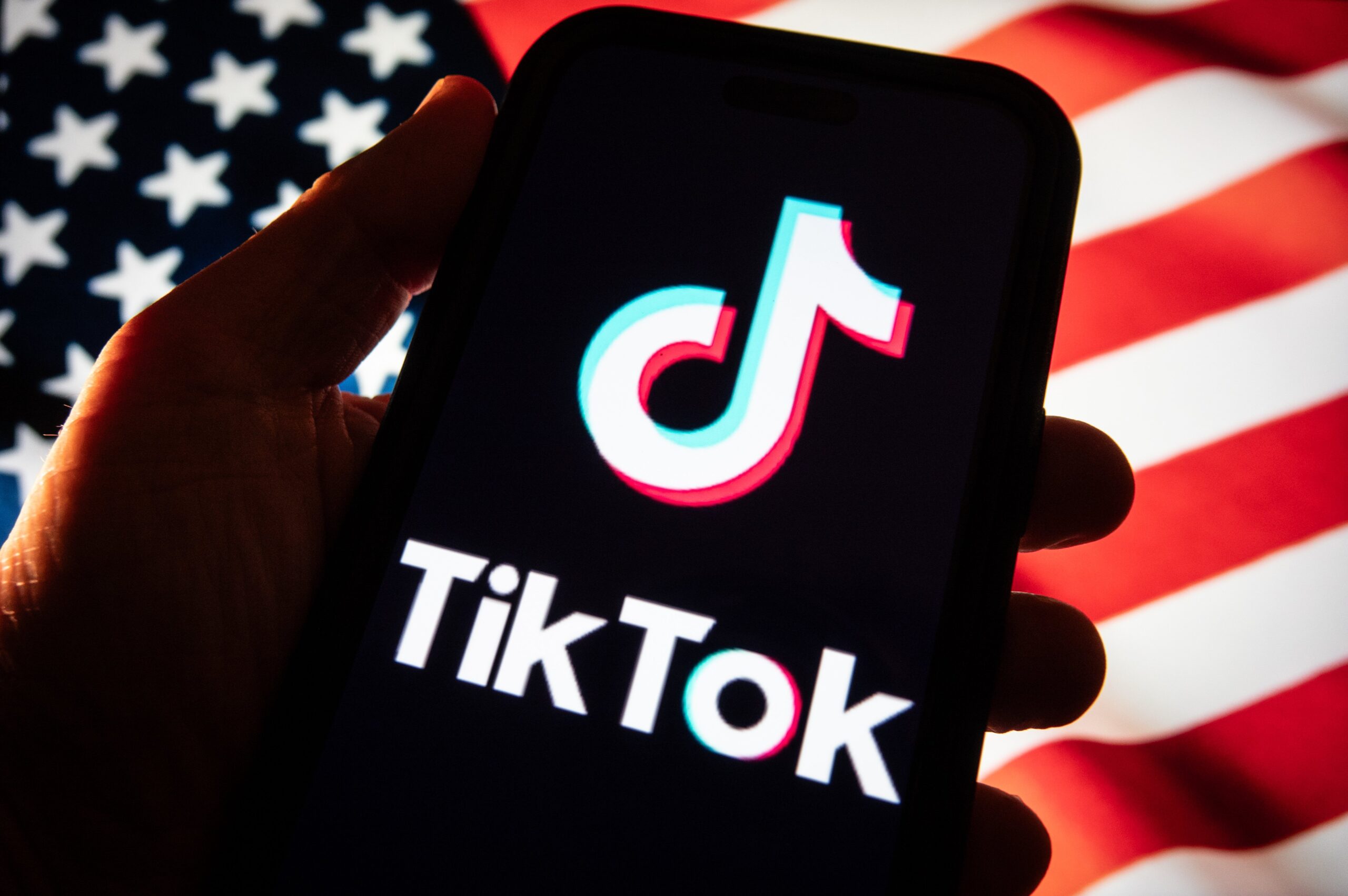 A hand holds out a phone with the TikTok logo in front of an American flag. 