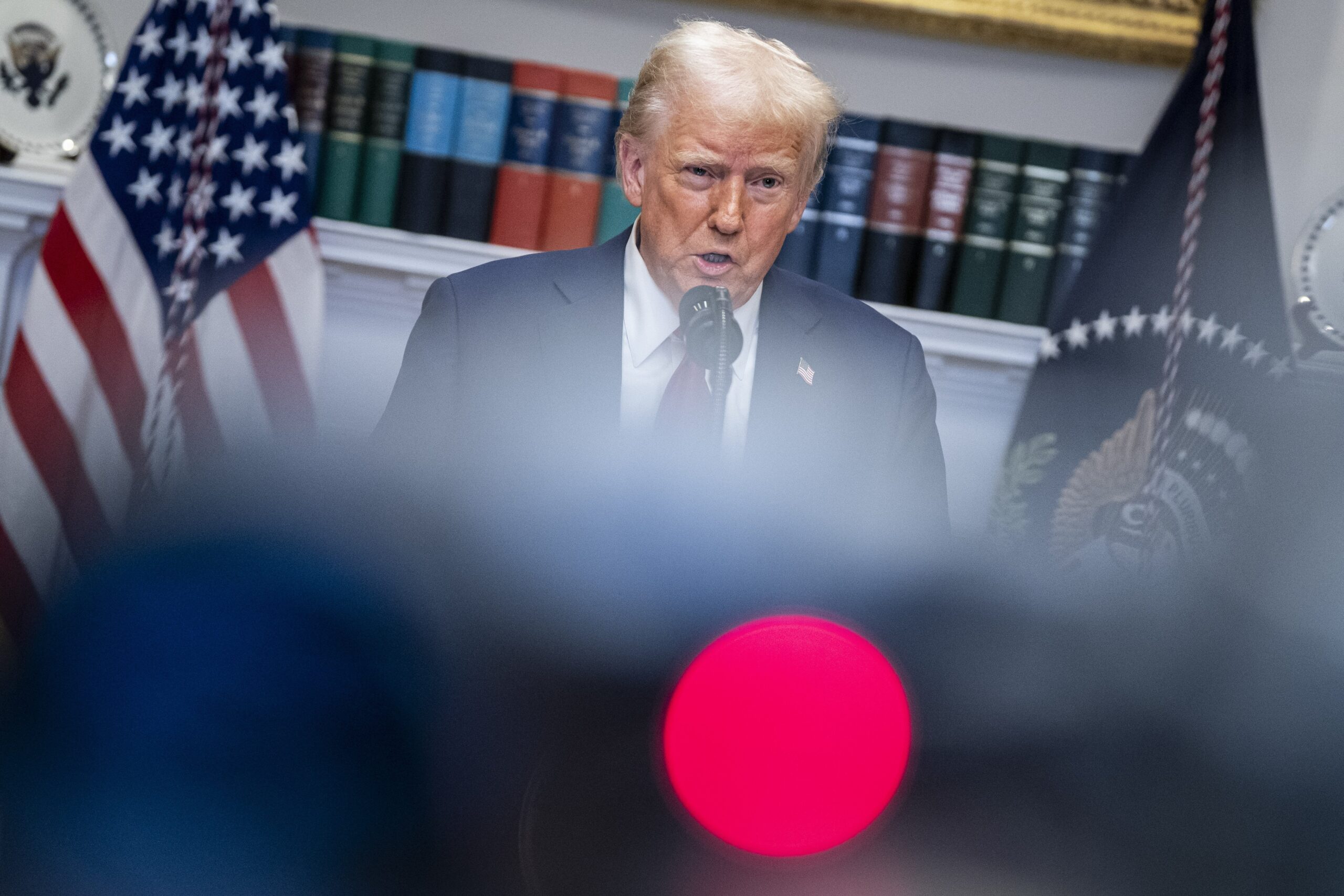 Trump stands at a podium surrounded by blurred out cameras and press members.