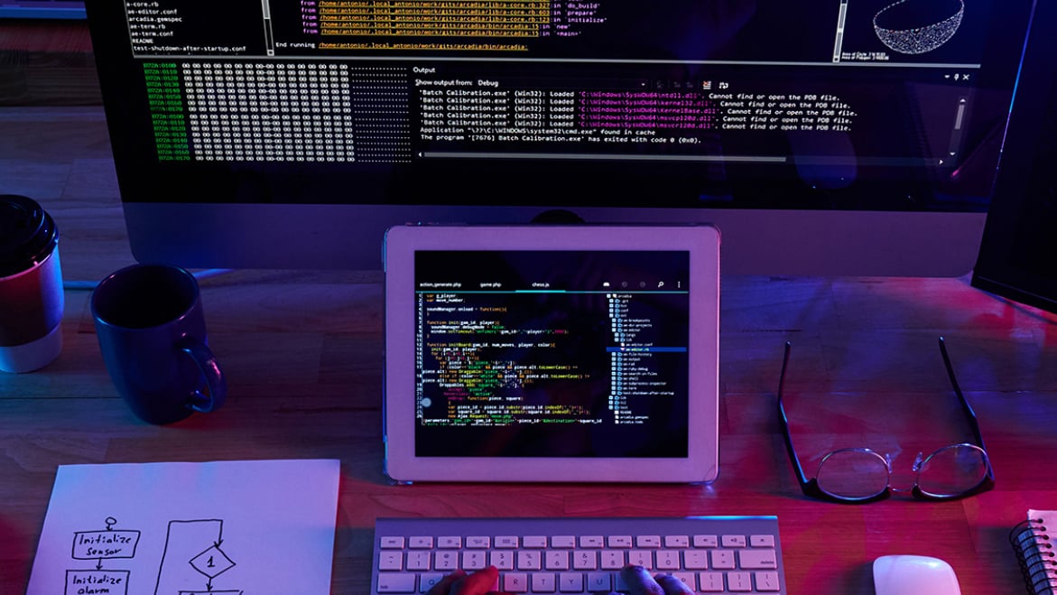 a crowded desk in a dark room with computer monitors showing programming language