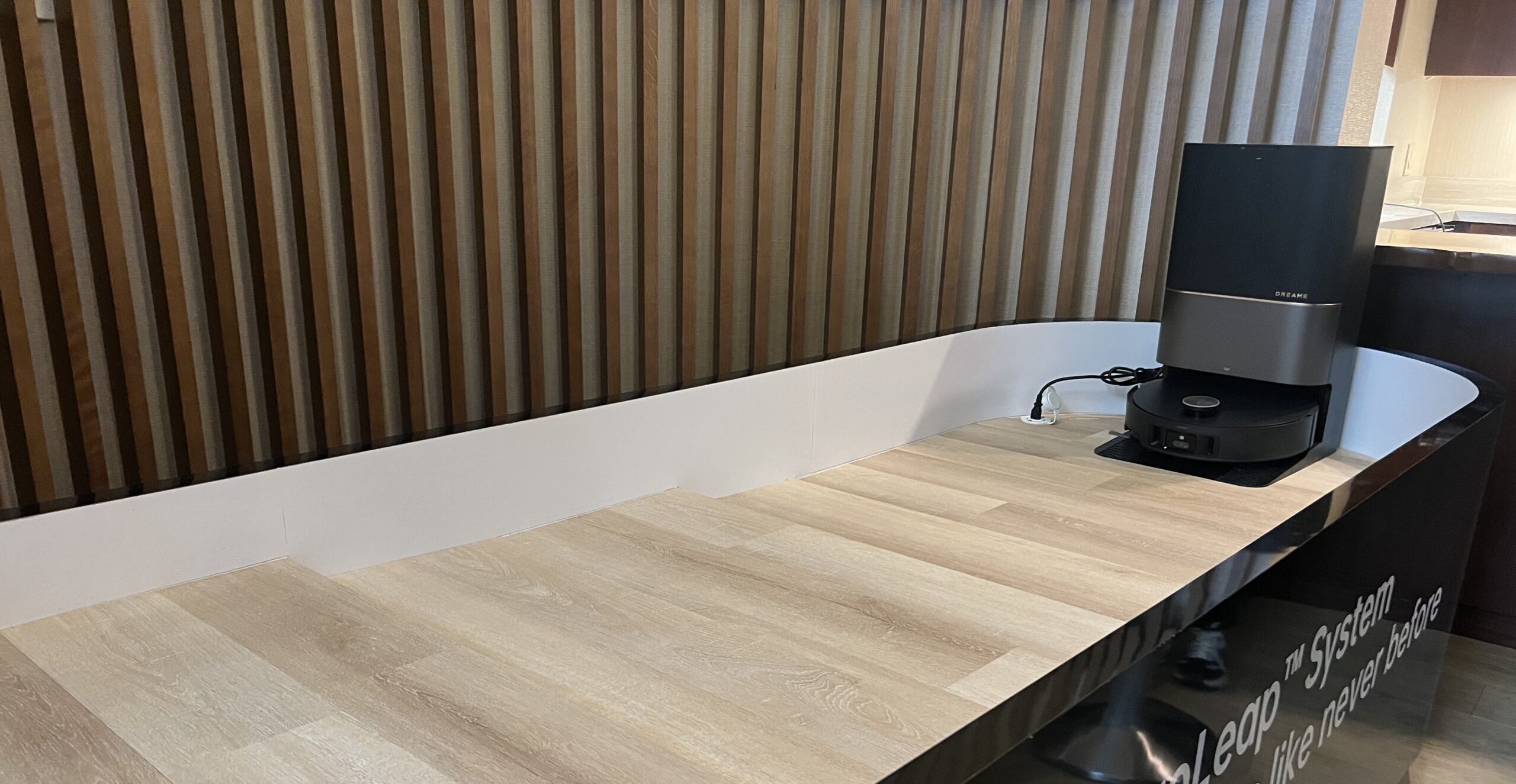 A robot vacuum and its charging dock displayed on a wooden platform with a white barrier and striped wooden wall background.