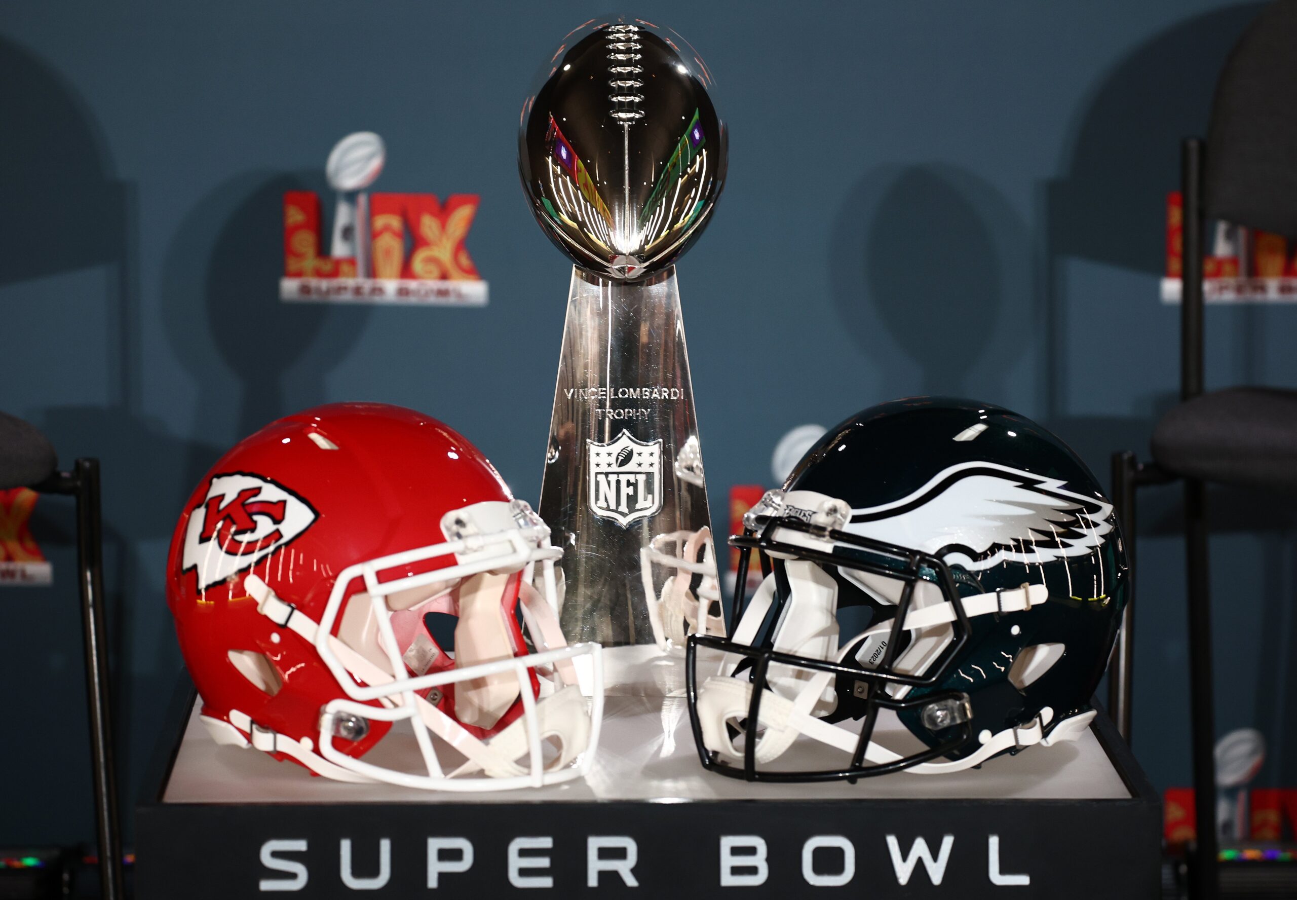 Two football helmets, one of with the Chiefs logo and the other with the Eagles, alongside the Super Bowl trophy on a table.