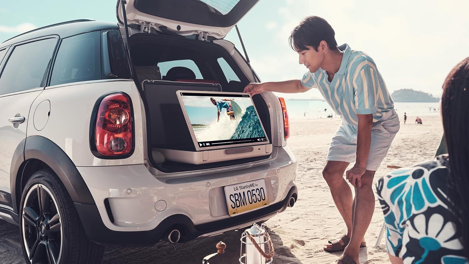 a person reaches to adjust the LG standbyme go portable monitor that's sitting in the open trunk of a car on a sunny beach