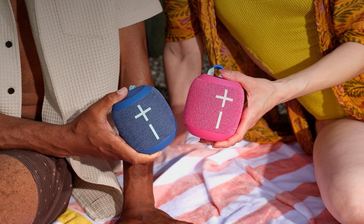 two people sitting on a beach towel each hold up an ultimate ears wonderboom 4 speaker. One speaker is blue and the other is pink.