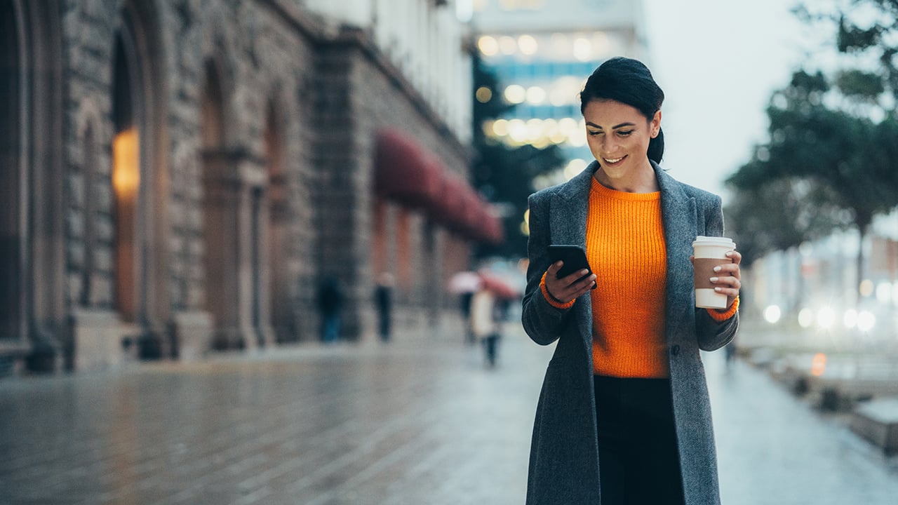 a woman walking on the street in a bright orange sweater looking at her phone in her left hand