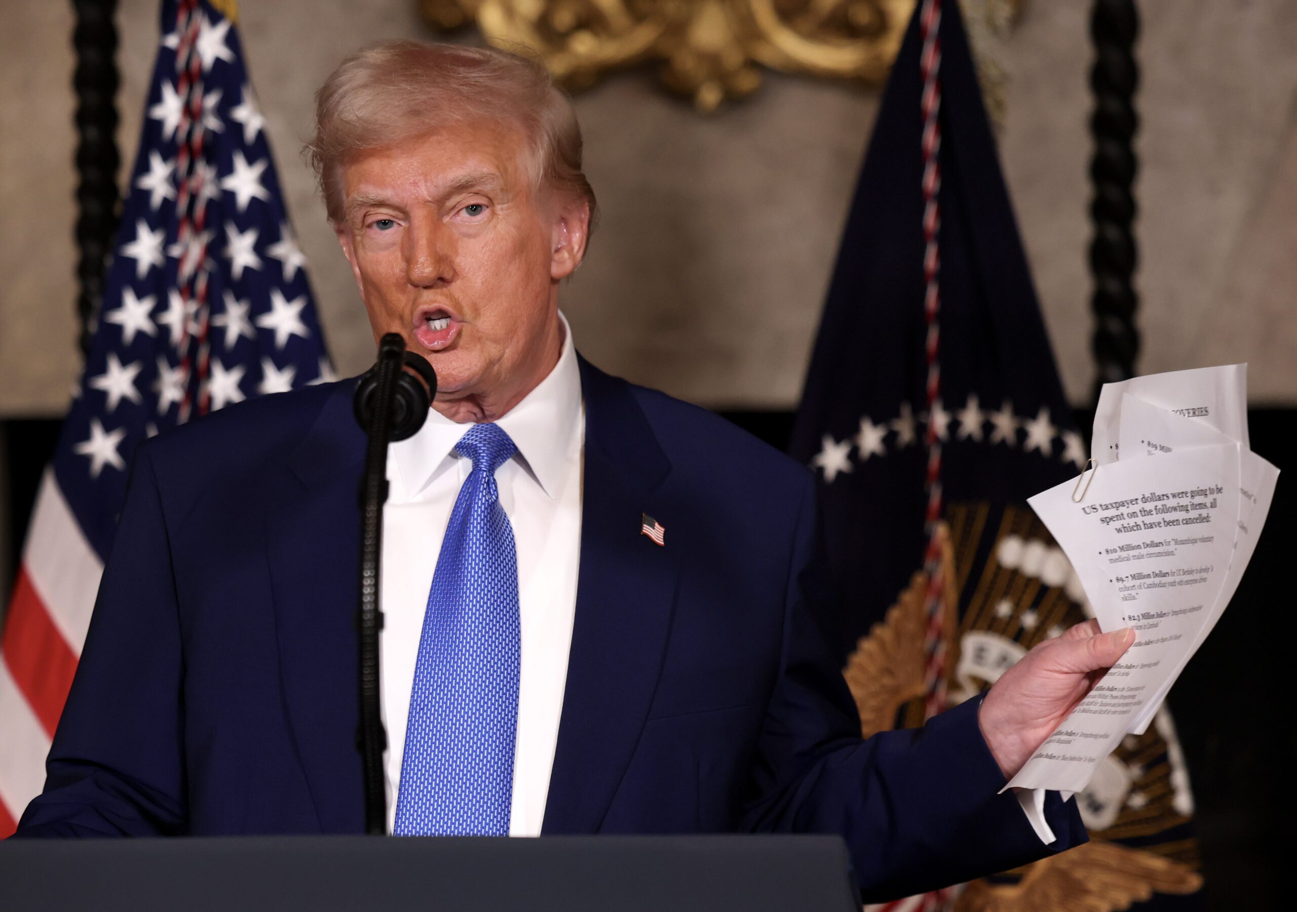 Trump holds up papers detailing DOGE spending cuts while speaking at a podium. 