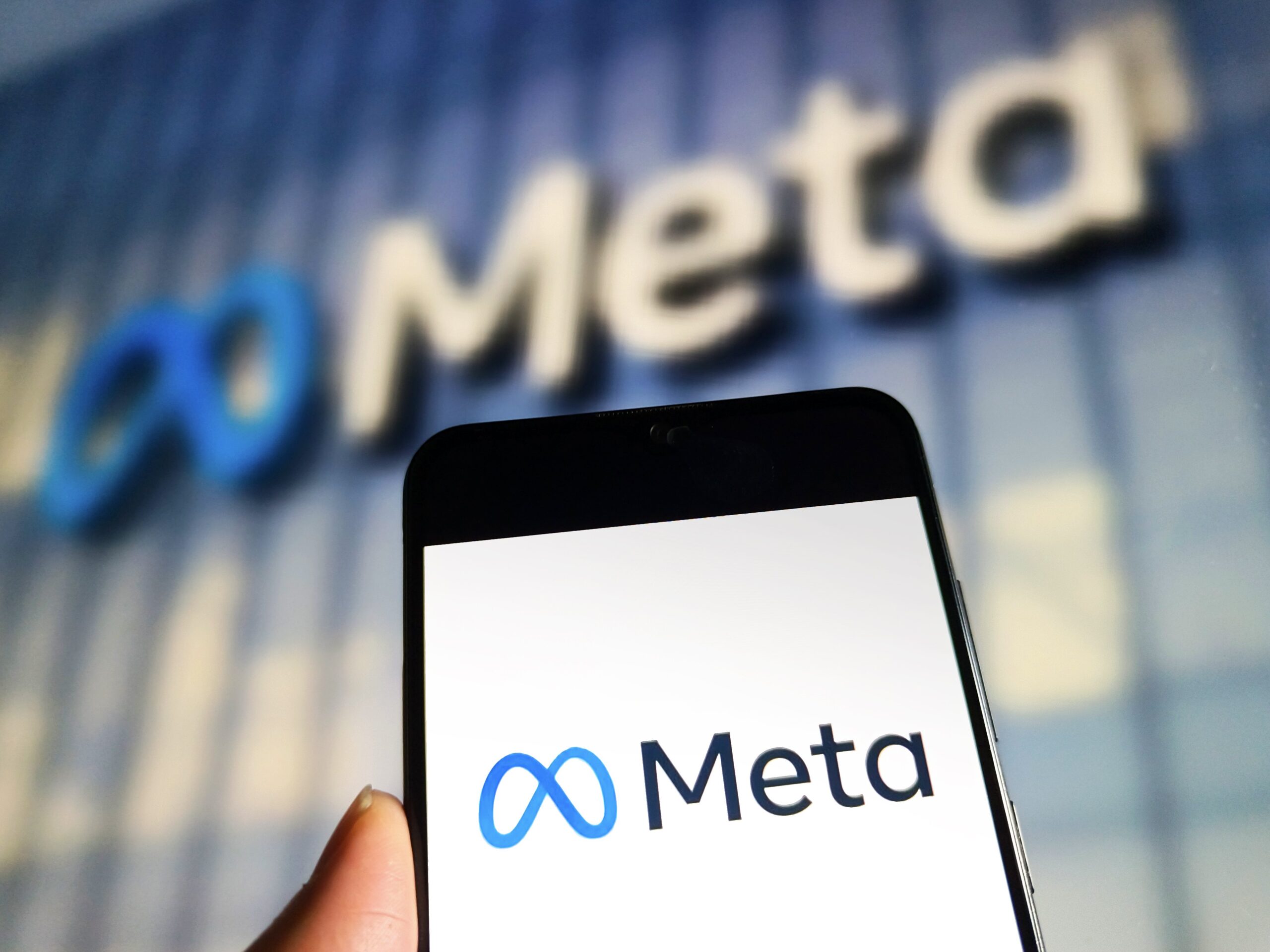 A phone displaying the Meta logo held up in front of a Meta office building.