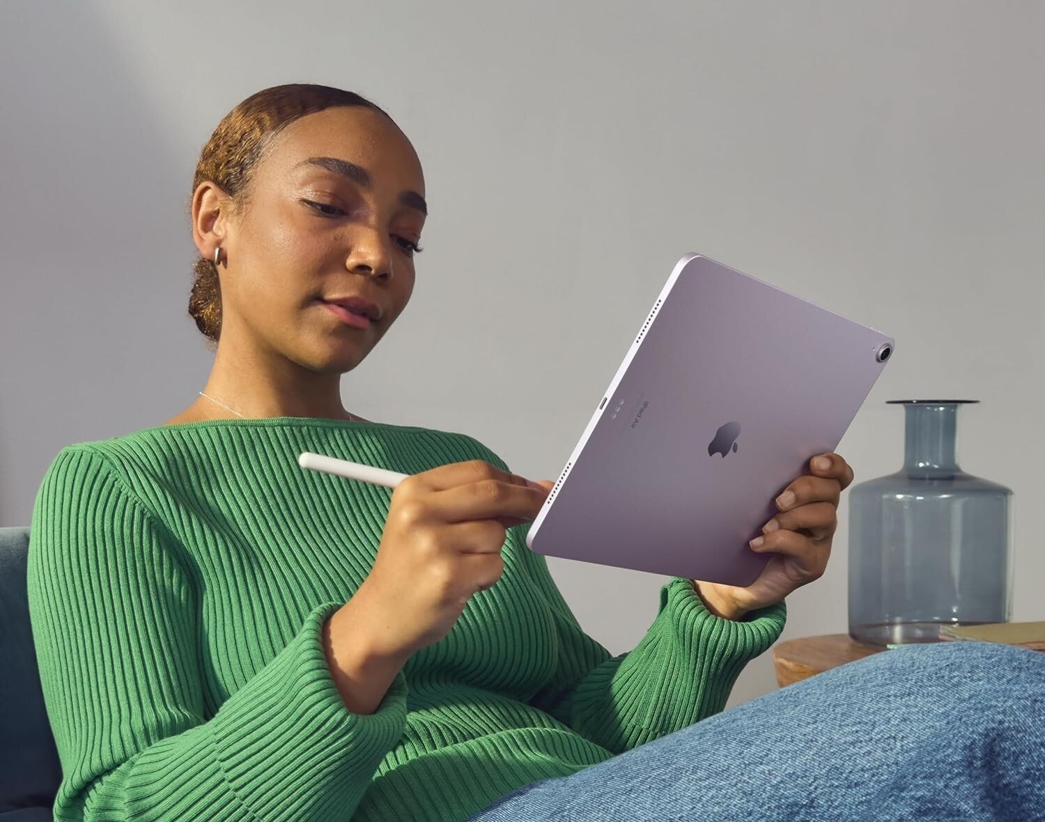 a person wearing jeans and a green sweater sits on a couch while using the apple ipad air with an apple pencil