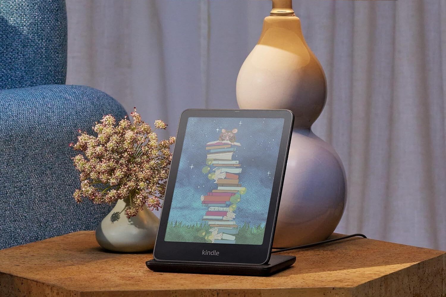 a kindle colorsoft e-reader sits on a side table on a stand with a lamp sitting behind it