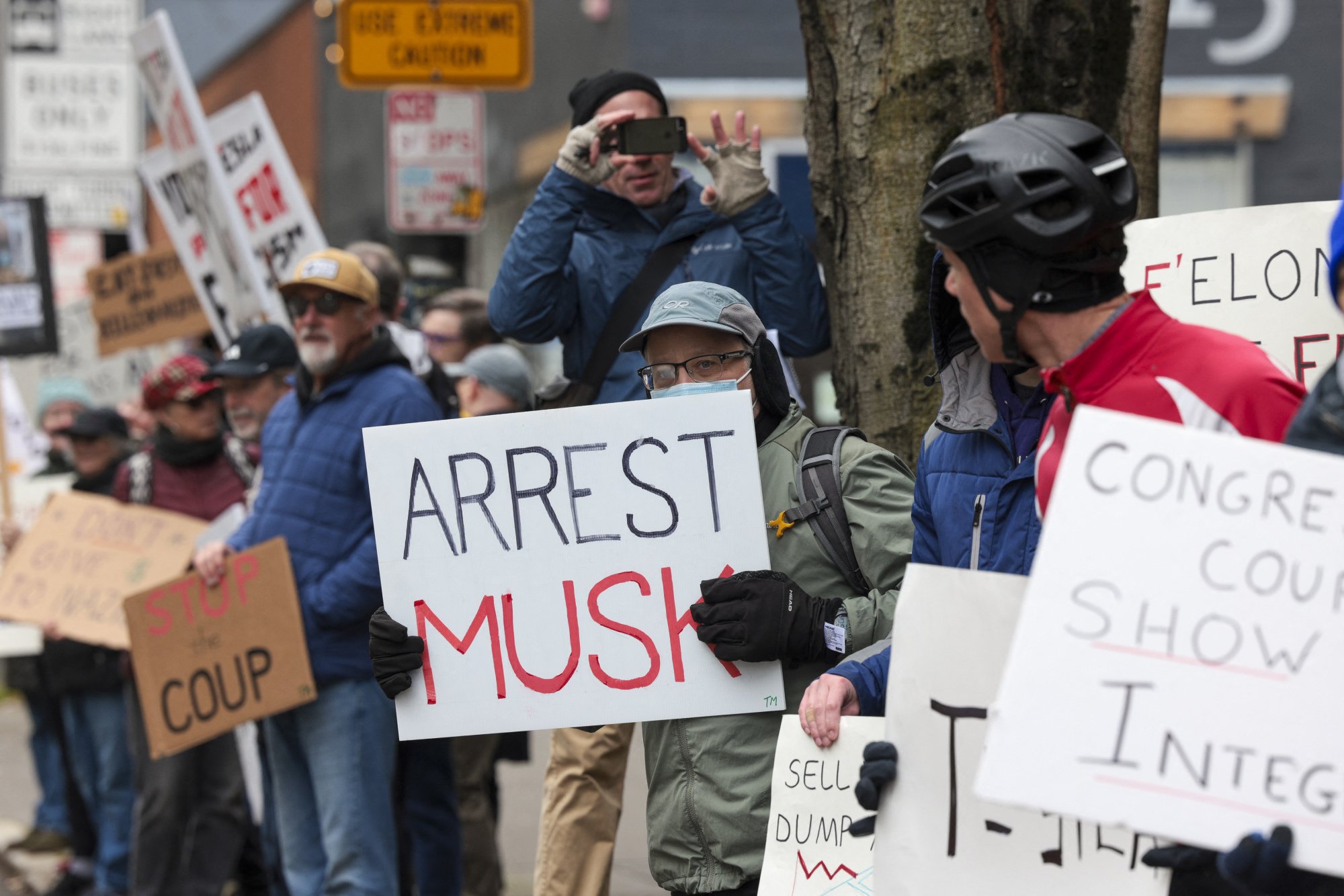 A group of protesters crowded together. One holds a sign that reads "Arrest Musk."
