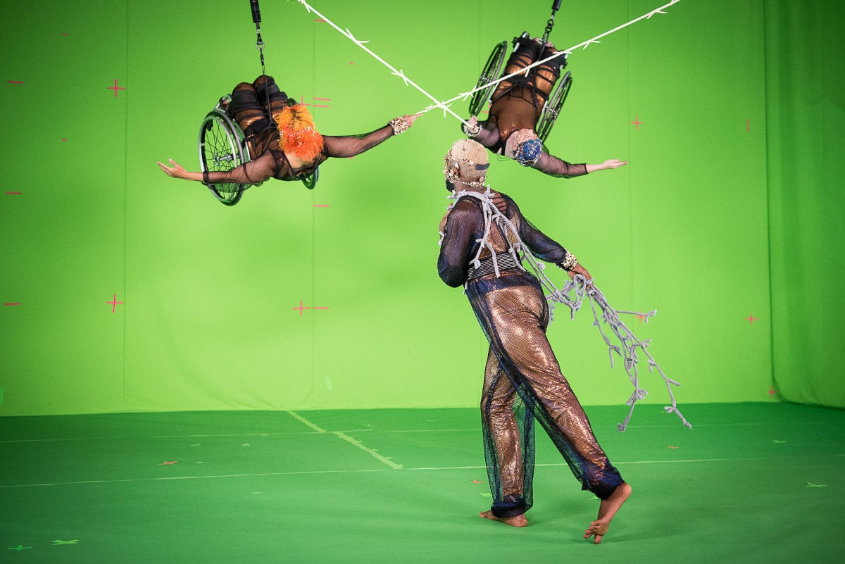 A behind-the-scenes moment from a film or performance set, featuring three performers in elaborate costumes against a green screen backdrop. Two of the performers are in wheelchairs, suspended in the air with harnesses, their arms outstretched toward each other. The third performer, standing barefoot on the ground, wears a netted outfit with decorative elements resembling vines or branches and appears to be interacting with the suspended performers. The green screen has visible tracking markers, indicating a visual effects setup.