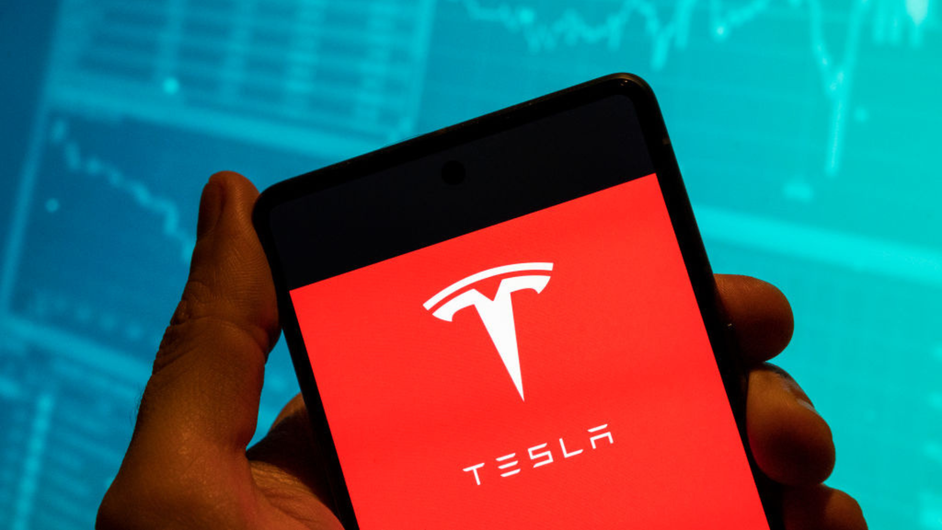 The Tesla logo is seen displayed on a smartphone with an economic stock exchange index graph in the background.