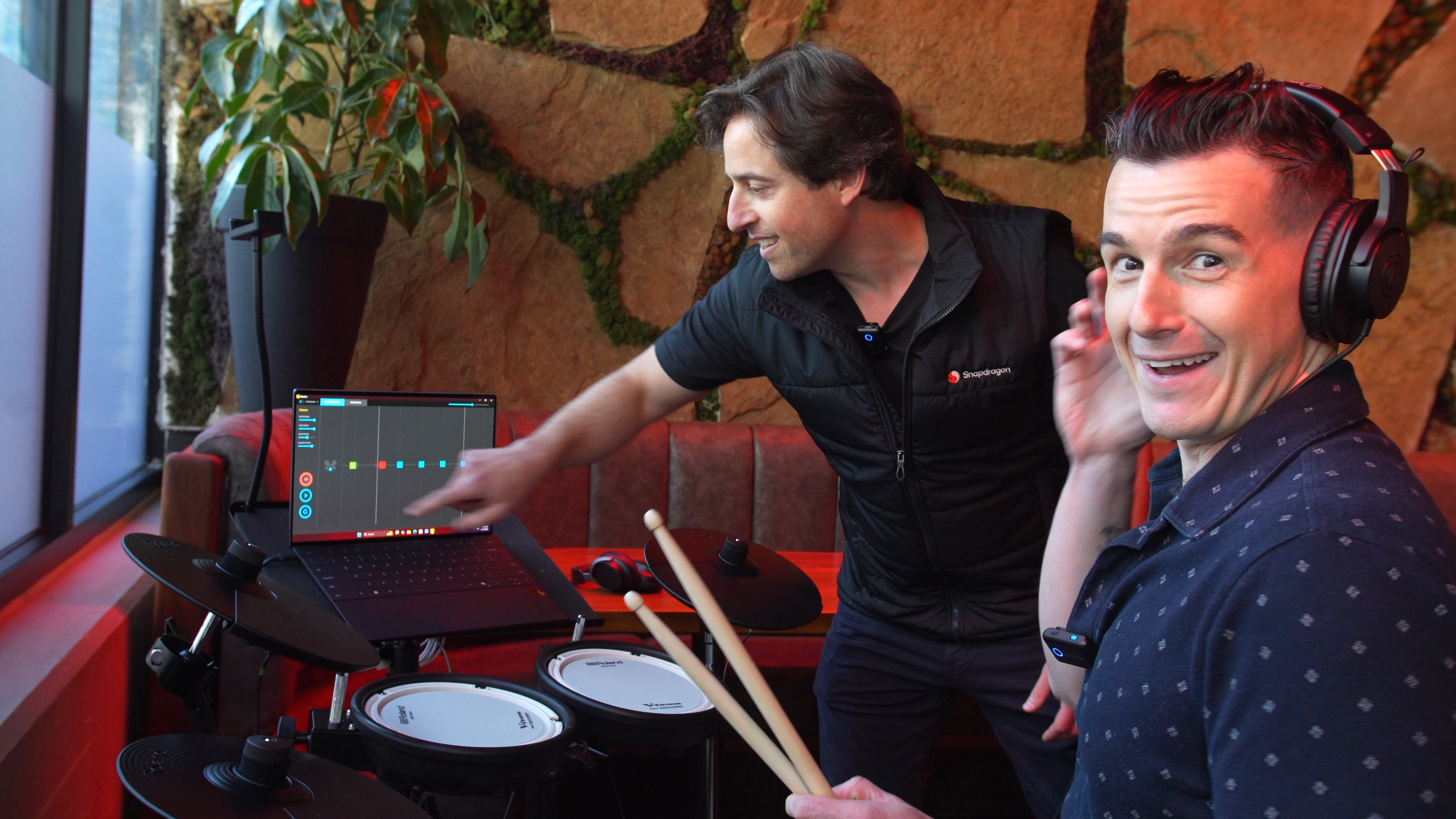 Man sitting at digital drum set while another man in the background adjusts music software on laptop