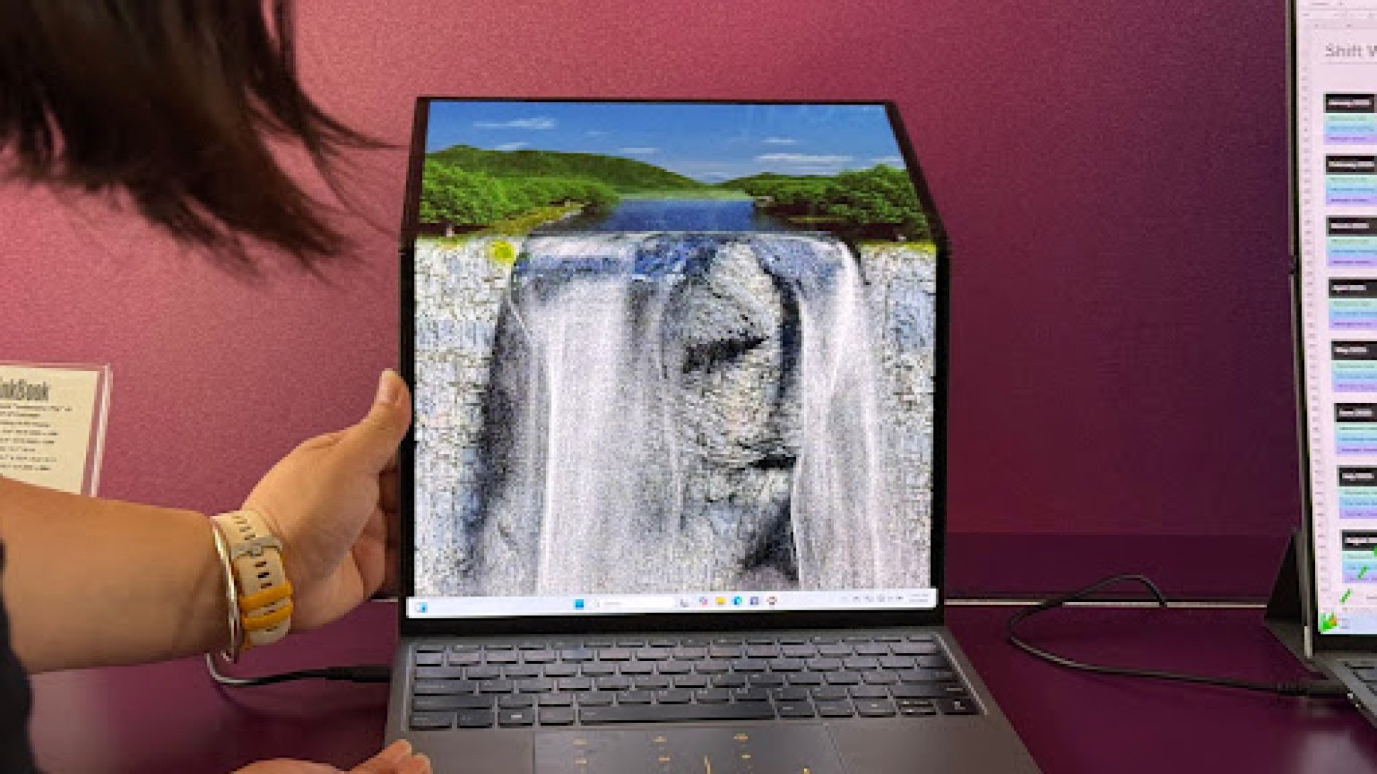 A person’s hand is adjusting a foldable laptop with a waterfall wallpaper, showing the device's flexibility.