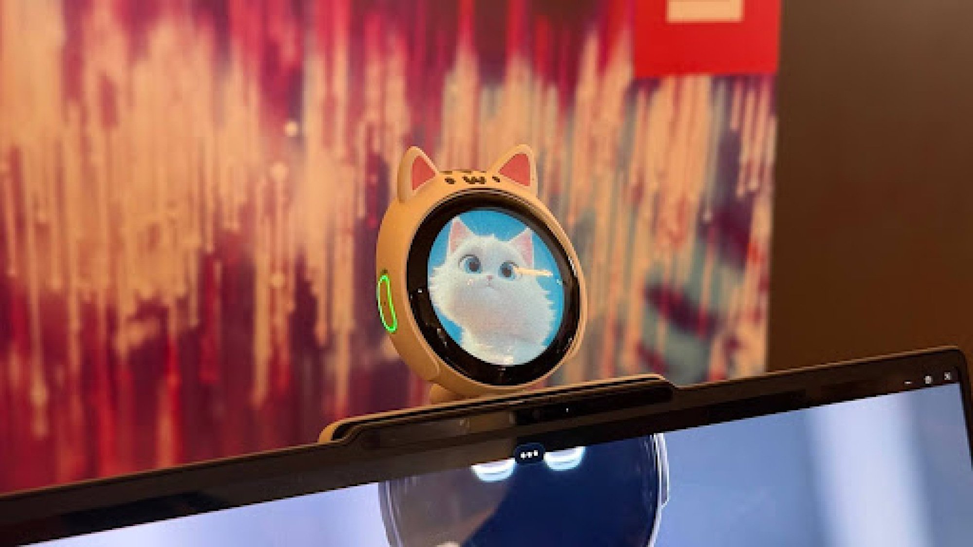 A round, cat-eared AI webcam with a digital pet face is perched on top of a computer monitor.