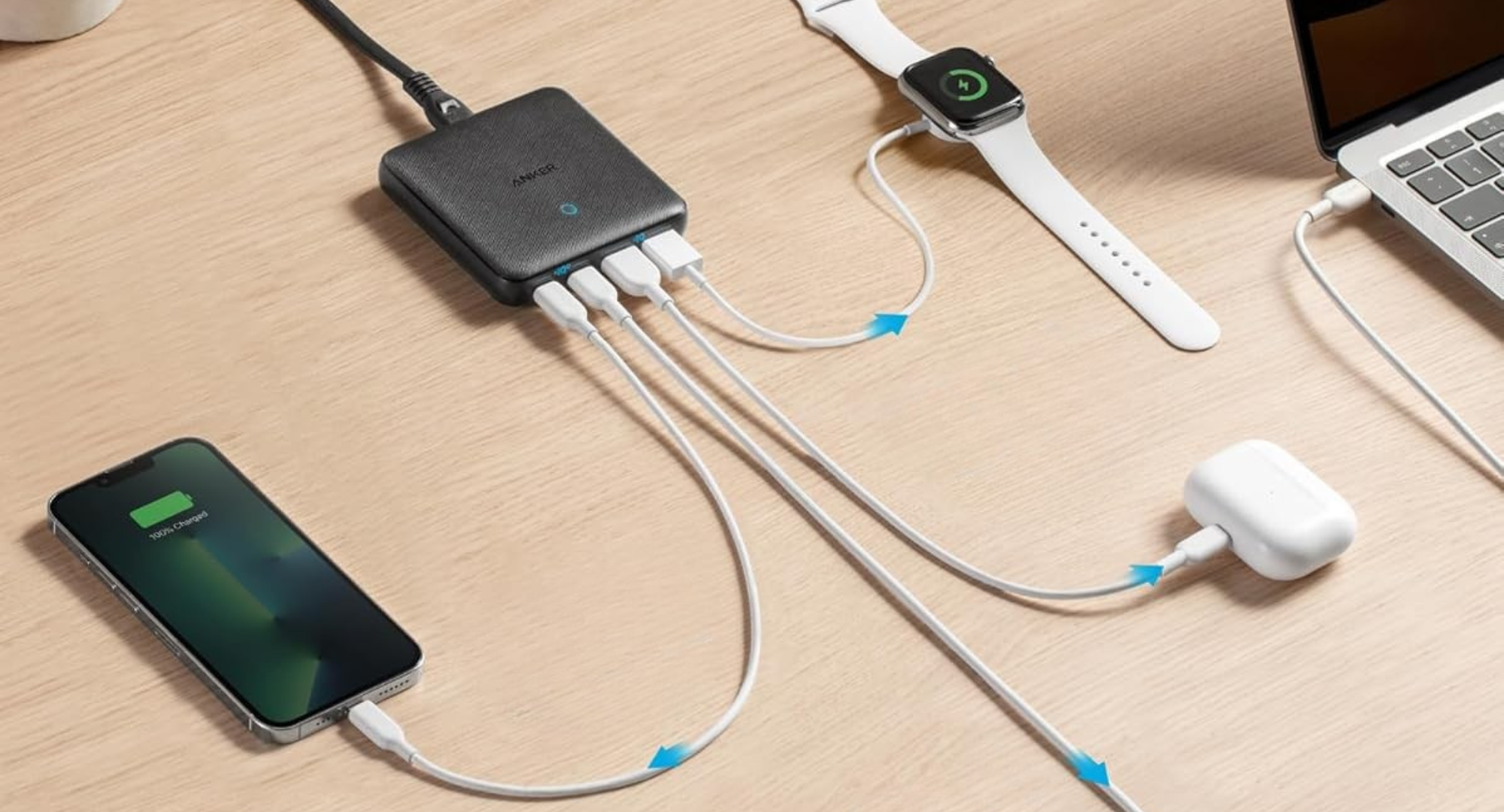 An Anker multi-port USB-C charging hub is shown connected to and charging a smartphone, Apple Watch, and AirPods. A laptop is visible in the background.