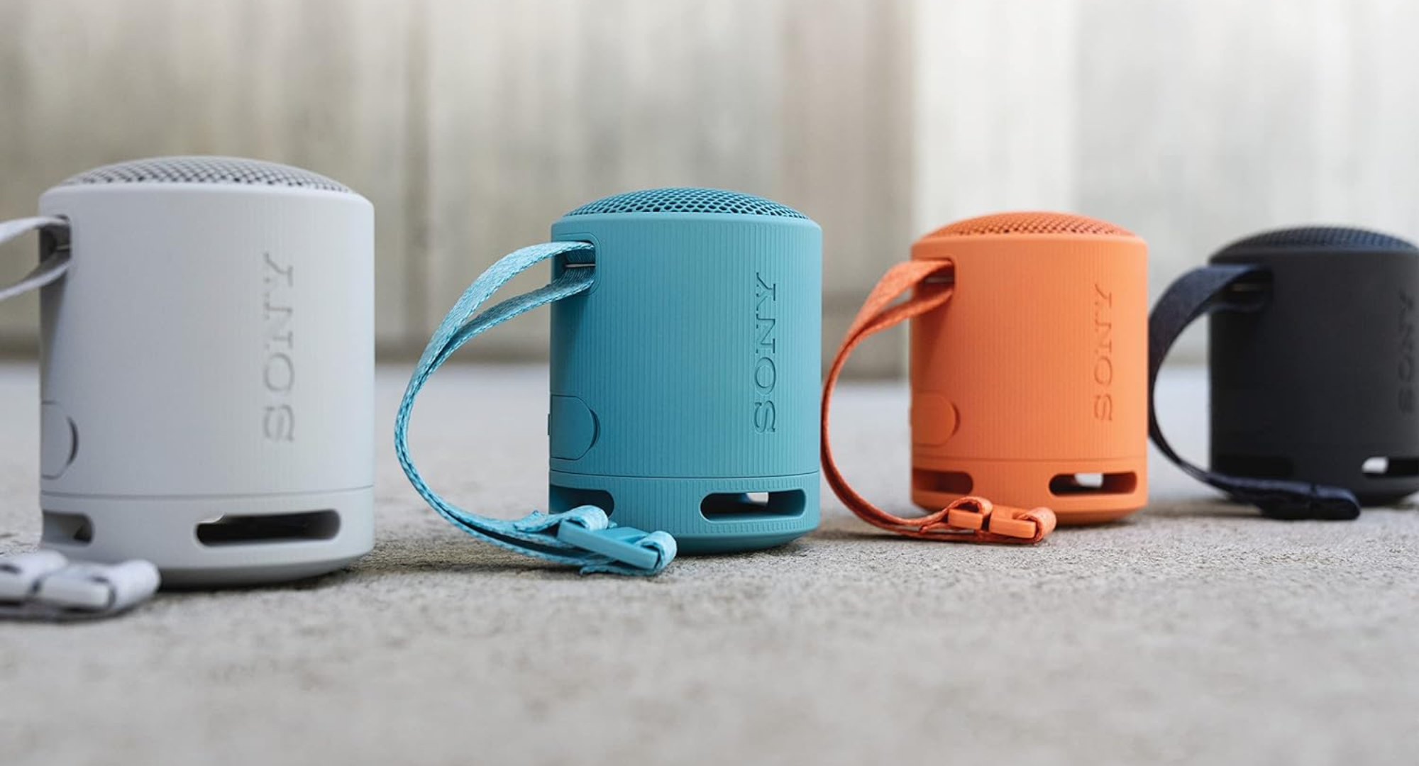 Four Sony SRS-XB100 portable speakers in light gray, blue, orange, and black are lined up next to each other, each with its carrying strap visible.