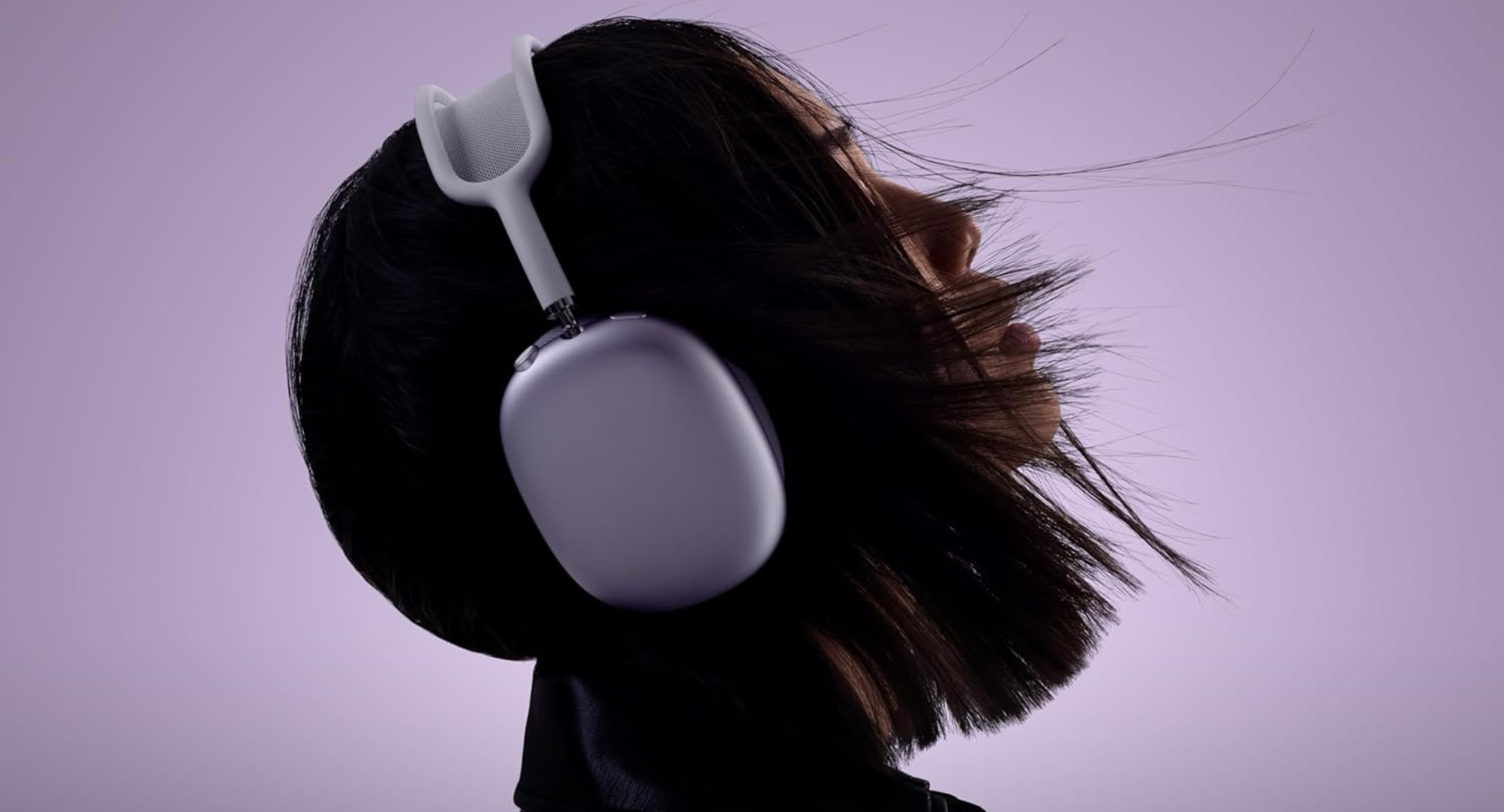 A person with dark, slightly windblown hair is shown from the shoulders up, facing slightly to the right. They are wearing white and silver Apple AirPods Max headphones with the right ear cup prominently displayed. The background is a soft, solid purple.