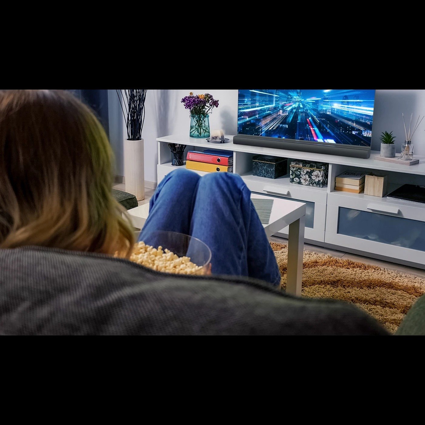 a person sits on the couch with a bowl of popcorn while watching TV. The Philips soundbar 2.0 sits below the TV