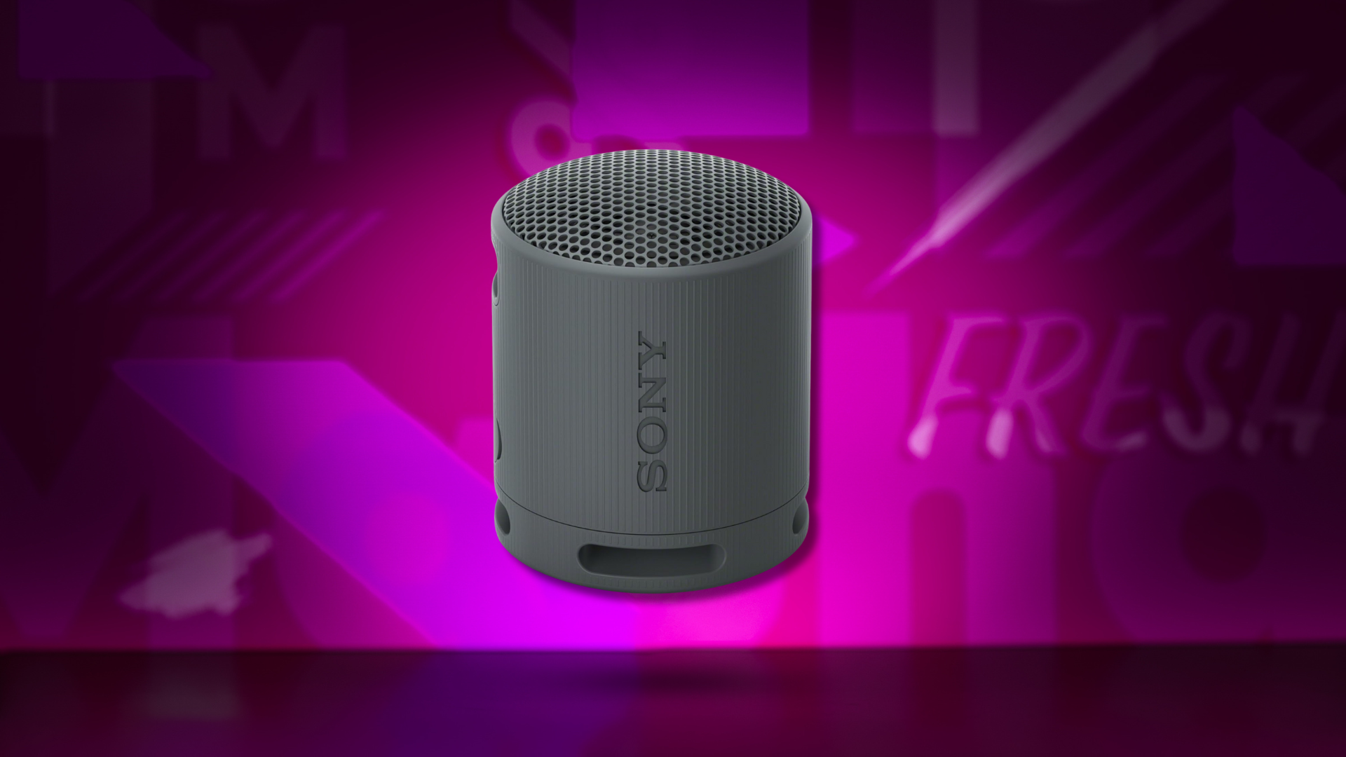 A small, cylindrical dark gray Sony SRS-XB100 portable Bluetooth speaker, standing upright with the 'SONY' logo visible on its side, set against a vibrant, blurred magenta and purple background with abstract text elements.