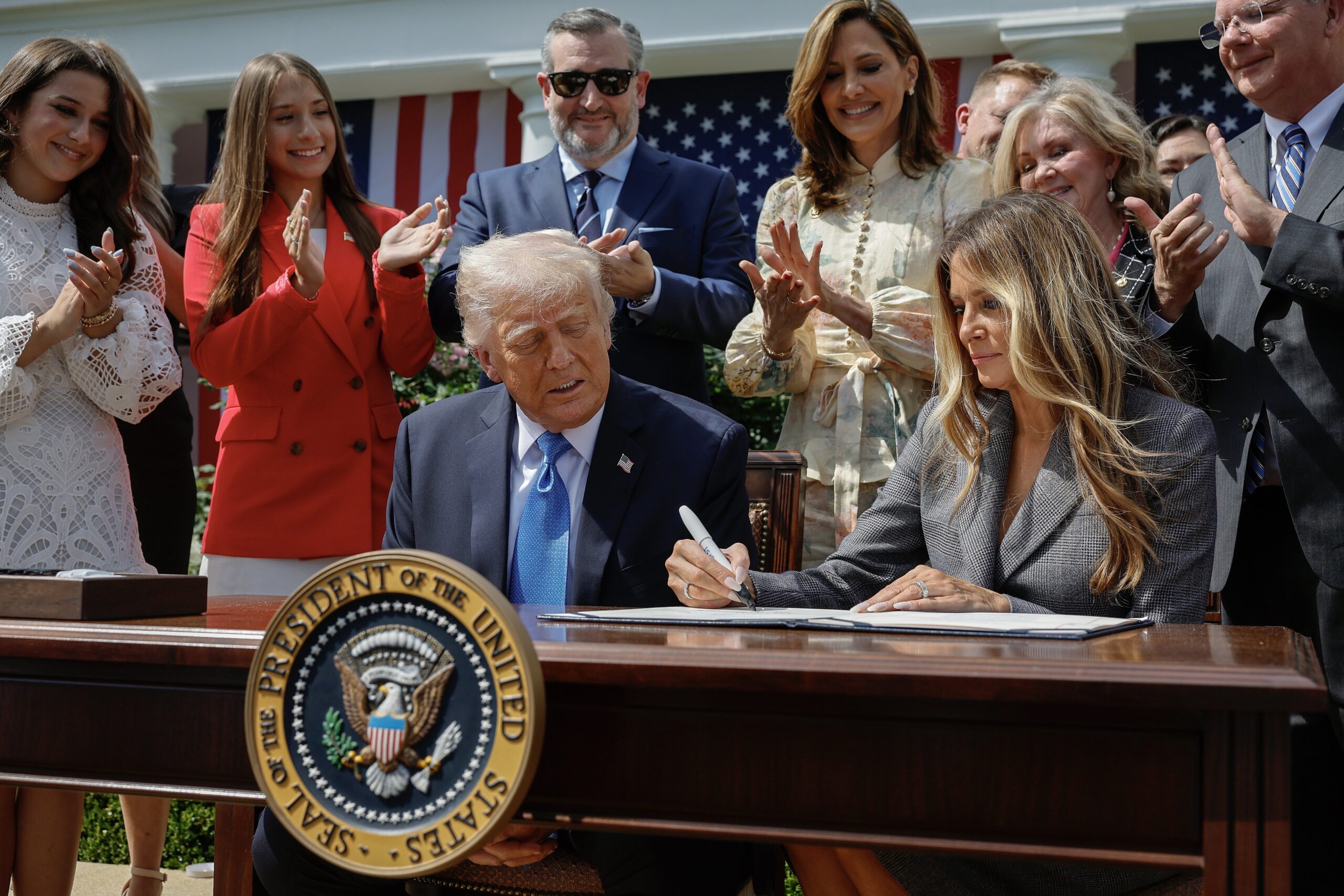 President Donald Trump and Melania Trump sit at a desk and sign the Take It Down Act while surrounded by people clapping.