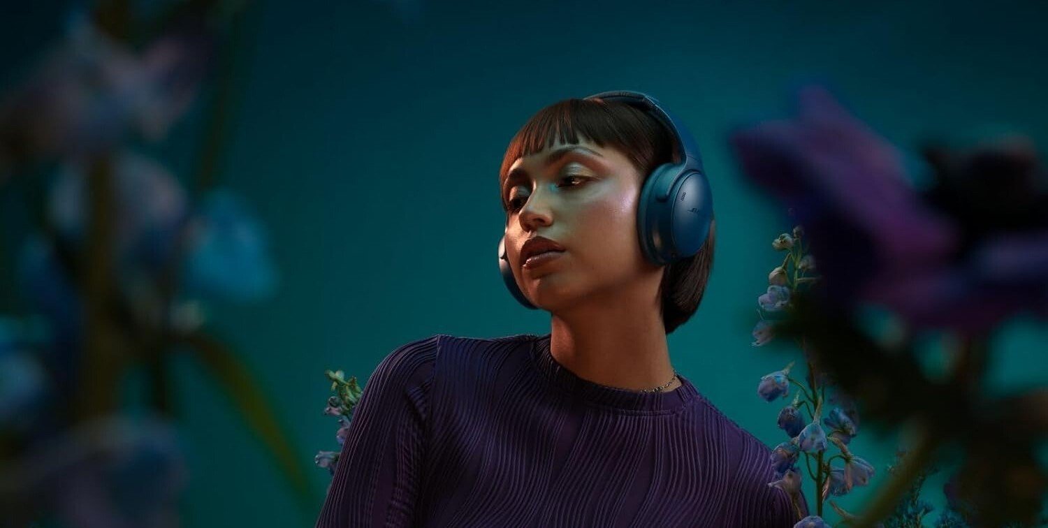 a person wears the bose QuietComfort headphone in the twilight blue colorway while standing near purple flowers with a purple backdrop