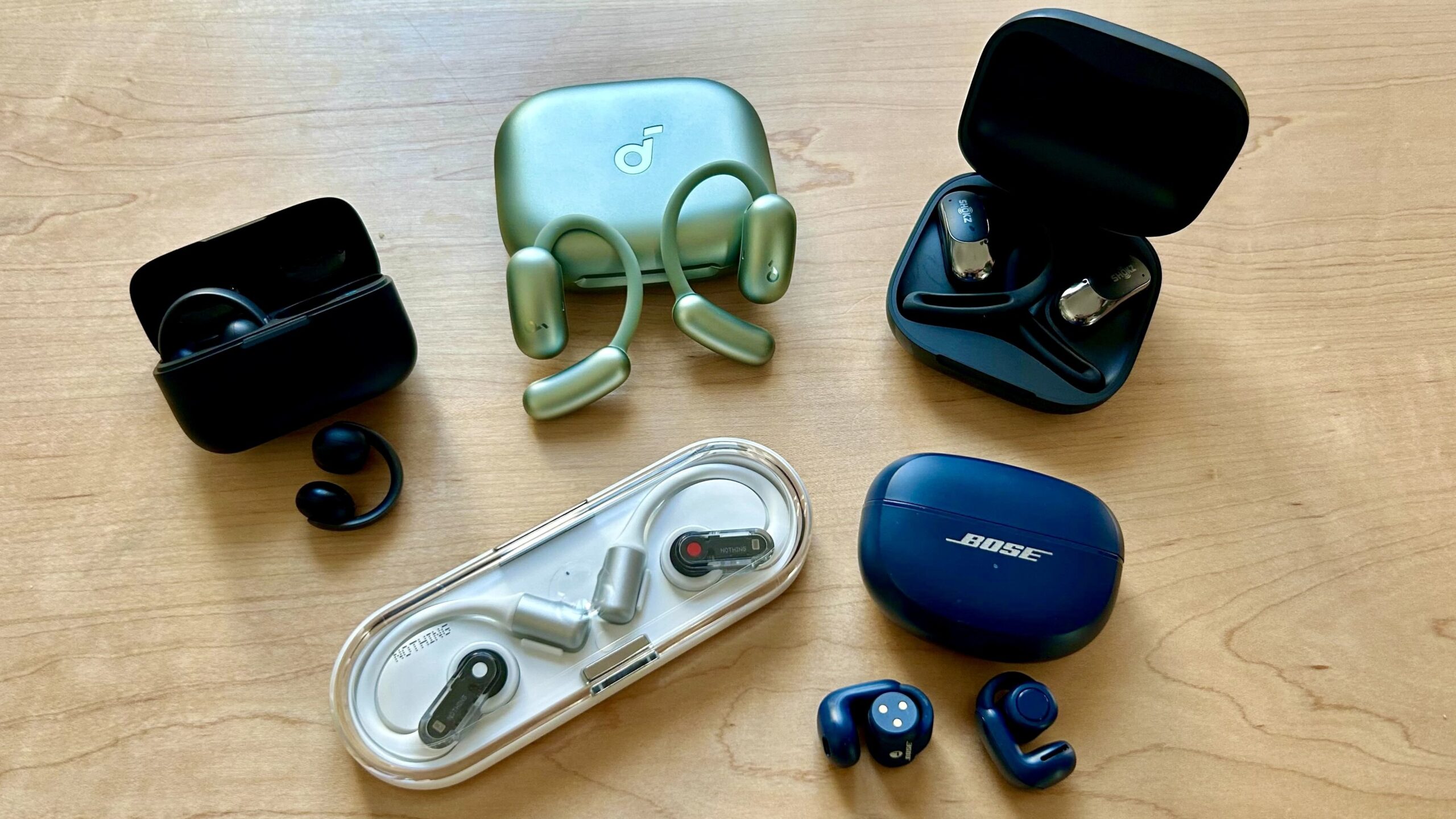 open earbuds from anker soundcore, shokz, nothing, and bose on oak tabletop