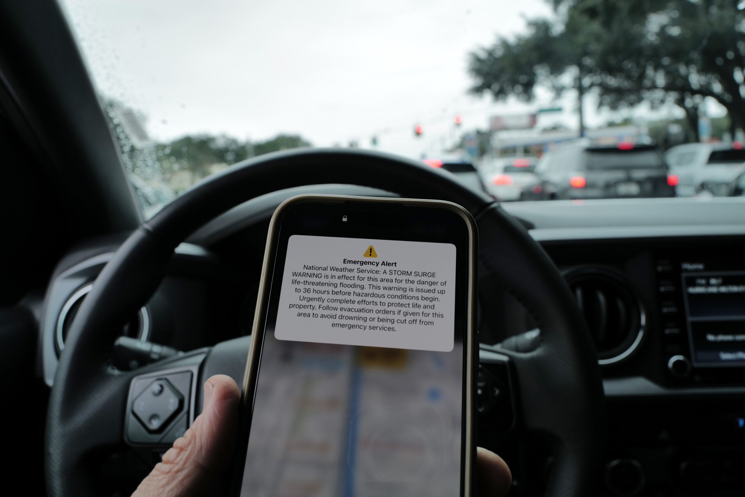 A hand holding an iPhone in front of a steering wheel. The phone shows an emergency alert notification for a storm surge.
