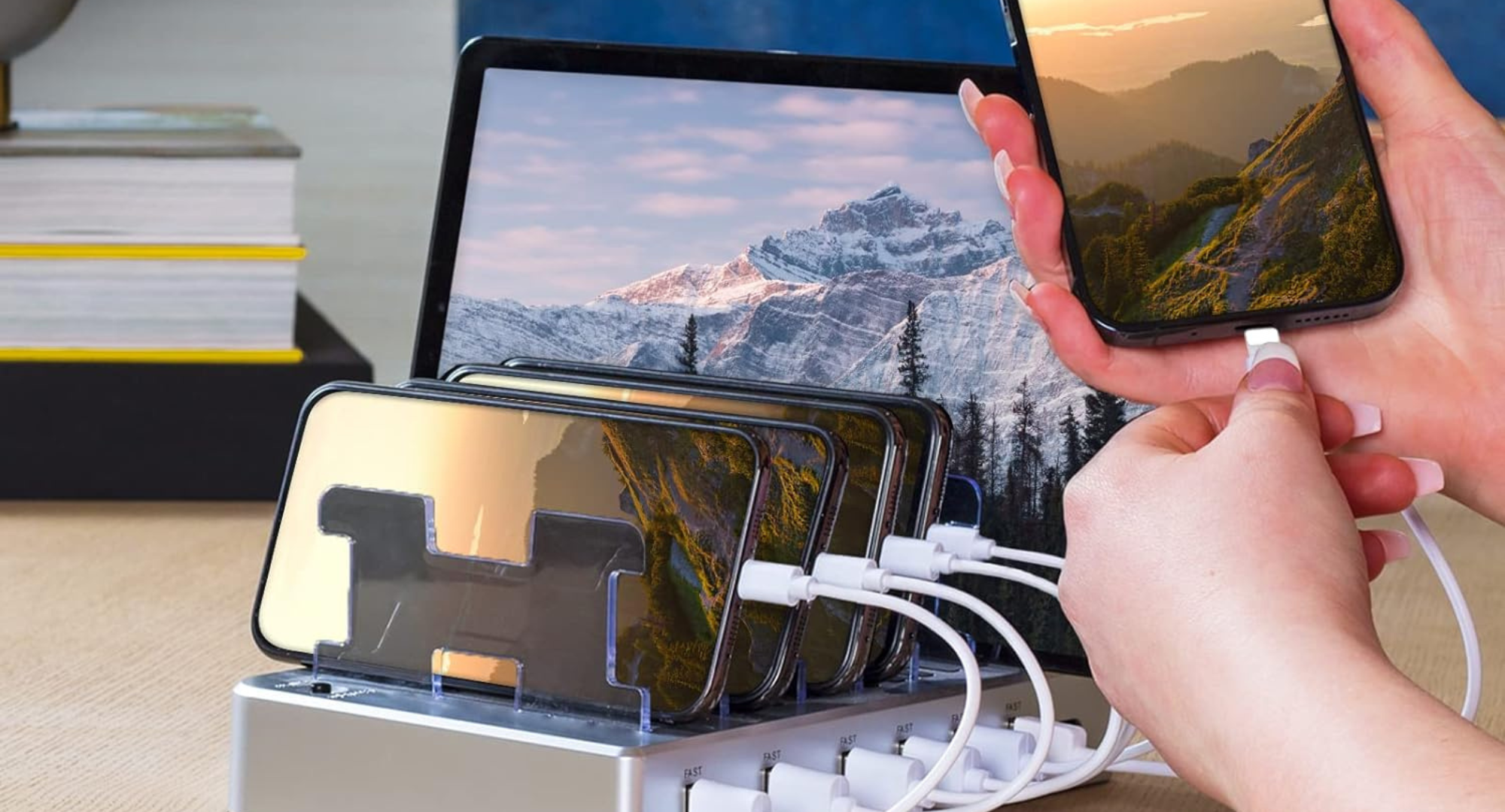 A silver 6-port charging station on a desk, simultaneously charging multiple smartphones and a tablet, with a hand plugging a cable into another smartphone.