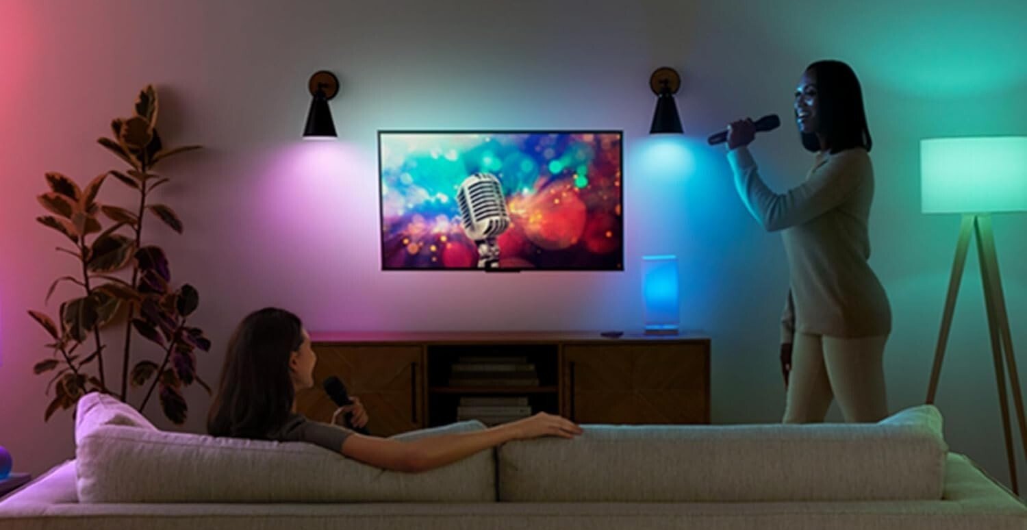 two people are in a dimly lit room aside from two amazon smart bulbs that are displaying purple and blue colors. One person is sitting on the couch and the other is standing while singing into a microphone