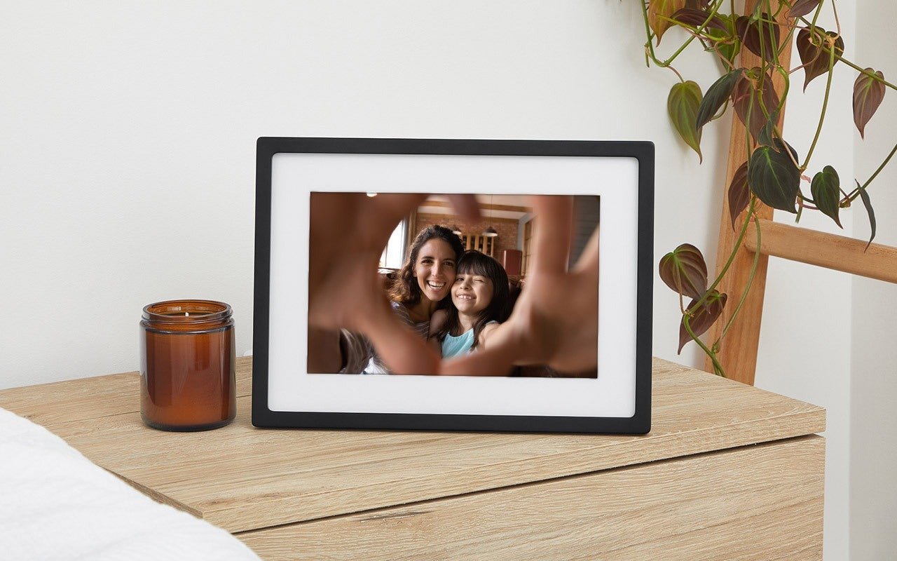 the skylight frame sits on a table with a plant hanging off the the right and a candle sitting to the left. the frame displays an image of a child and a woman sitting together and smiling