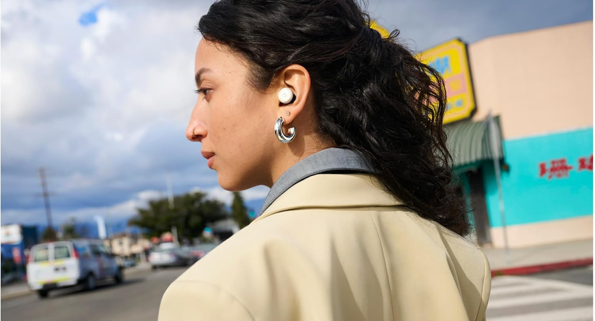 A person is shown from the side, looking to their left. They are wearing a white Google Pixel Bud Pro in their visible ear and a light-colored jacket. The background is slightly blurred, showing an outdoor street scene with cars and a building with some signage.