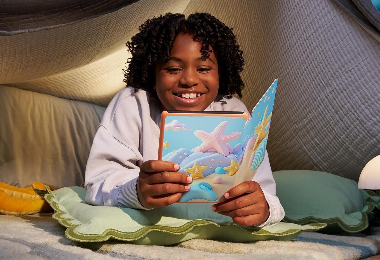 a child with curly hair lays in a pillow fort while reading on a kindle paperwhite kids and smiling