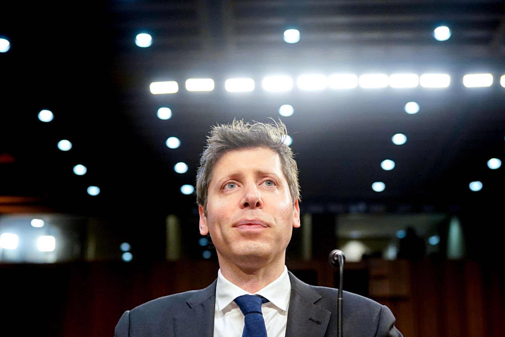 a closeup up Sam Altman wearing a suit with bright lights behind him