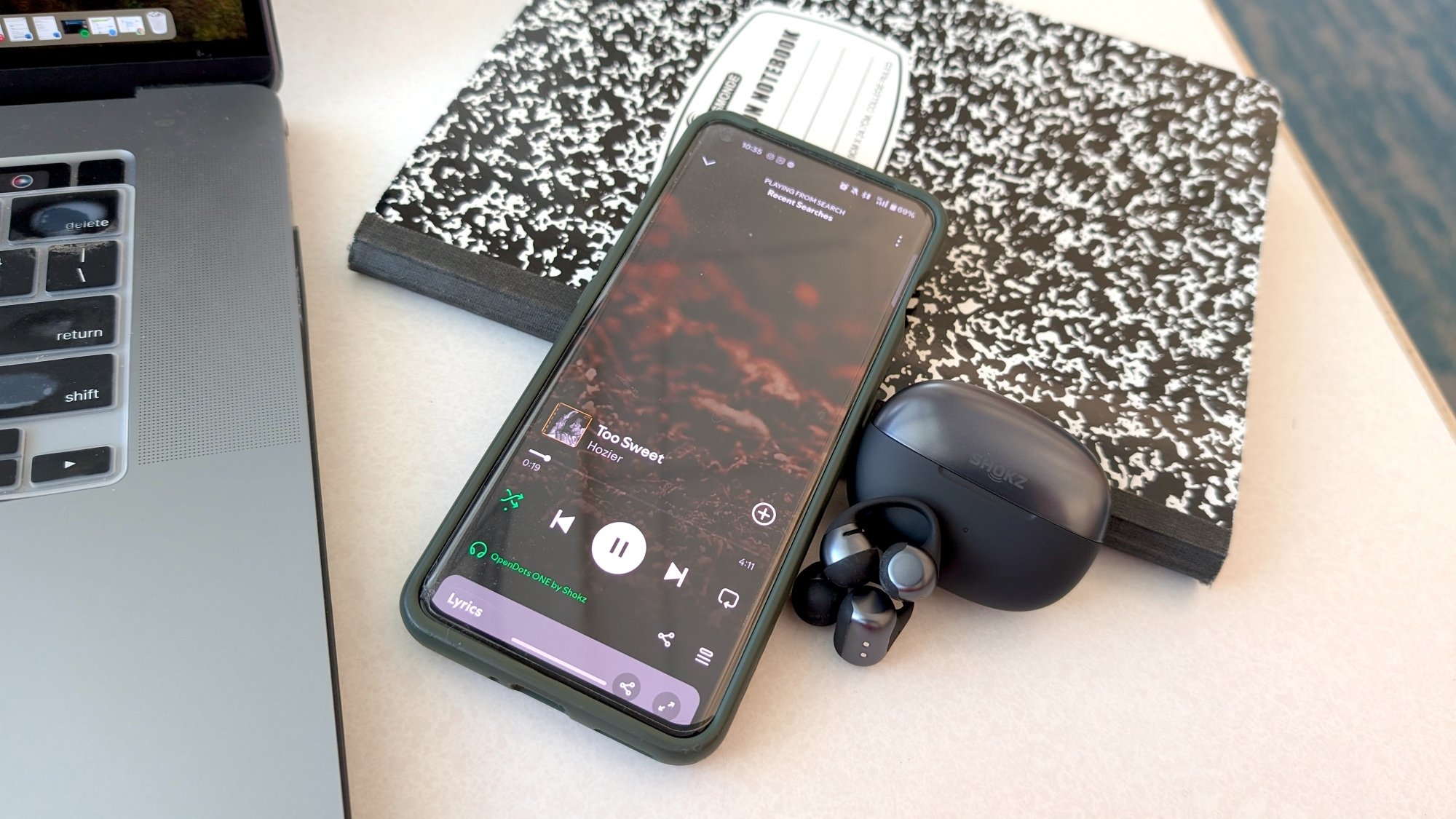 shokz opendots one earbuds next to phone with Hozer's Too Sweet on screen