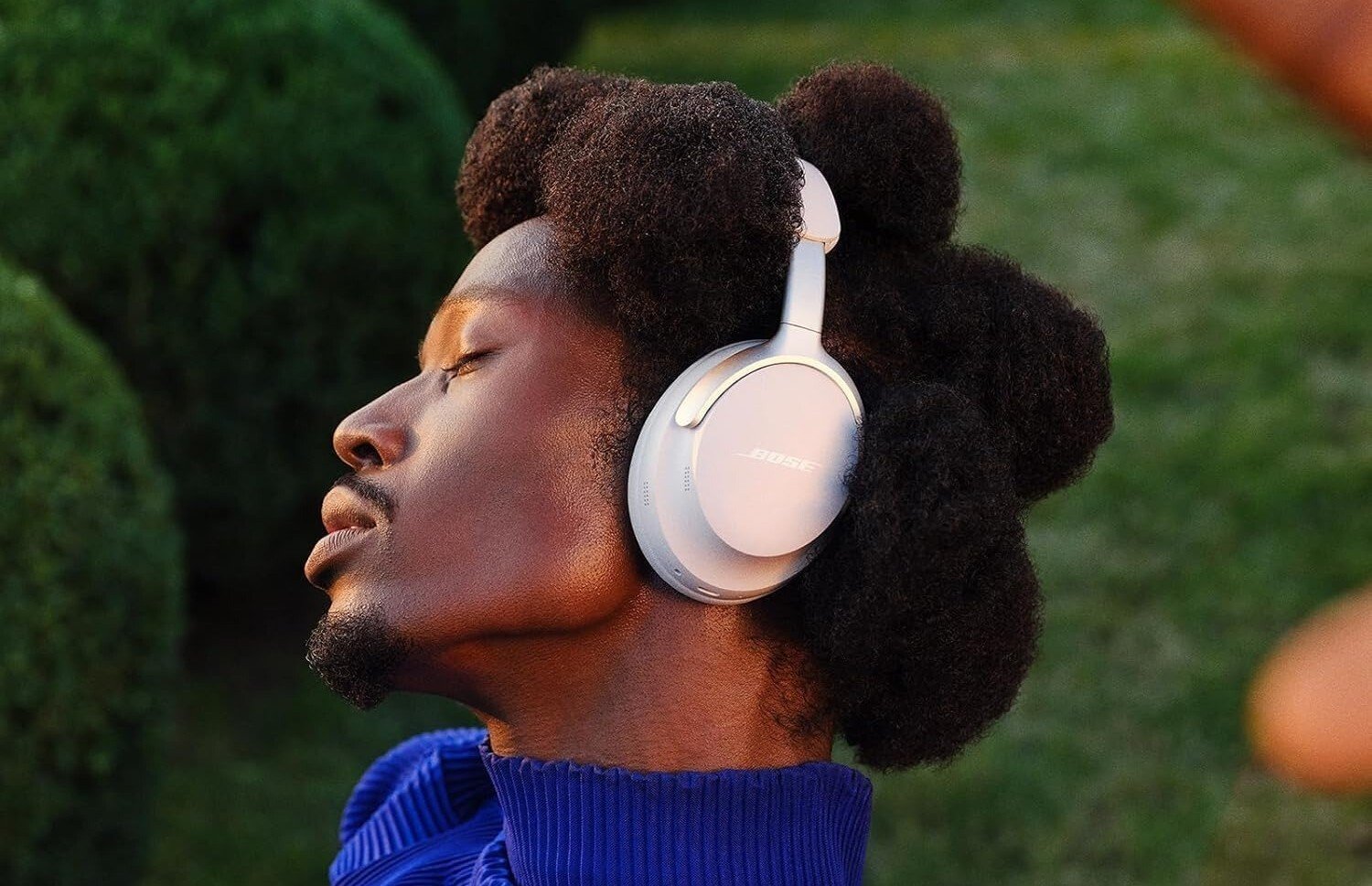 a person wears a white pair of bose quietcomfort ultra while closing their eyes. behind them is a lot of green bushes.
