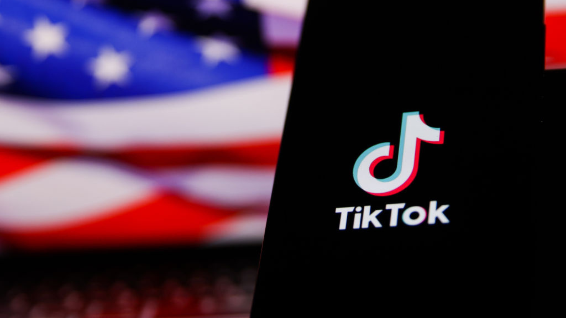 The TikTok logo on a mobile phone screen with the U.S. flag in the background.