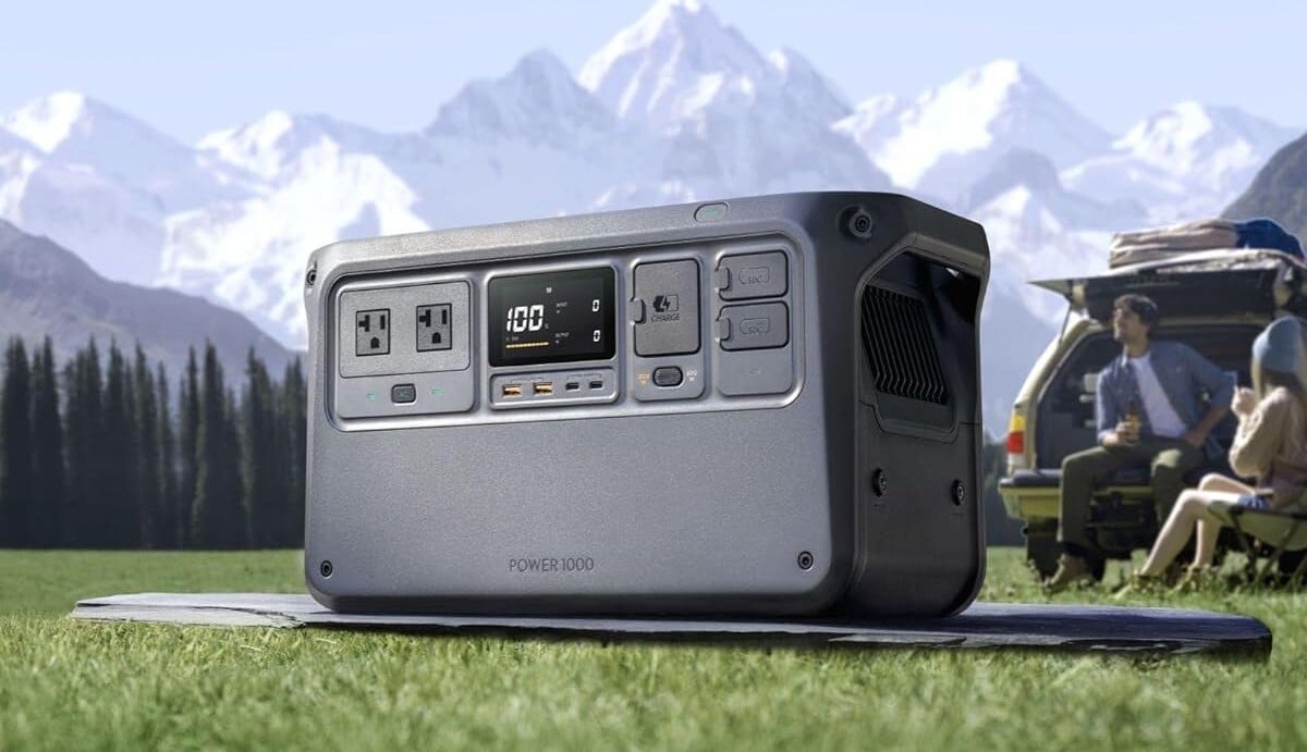 the DJI power 1000 portable power stations sits in a field of grass with a snowy mountain range behind on a sunny day. two people are sitting near the open trunk of a car that's parked nearby.