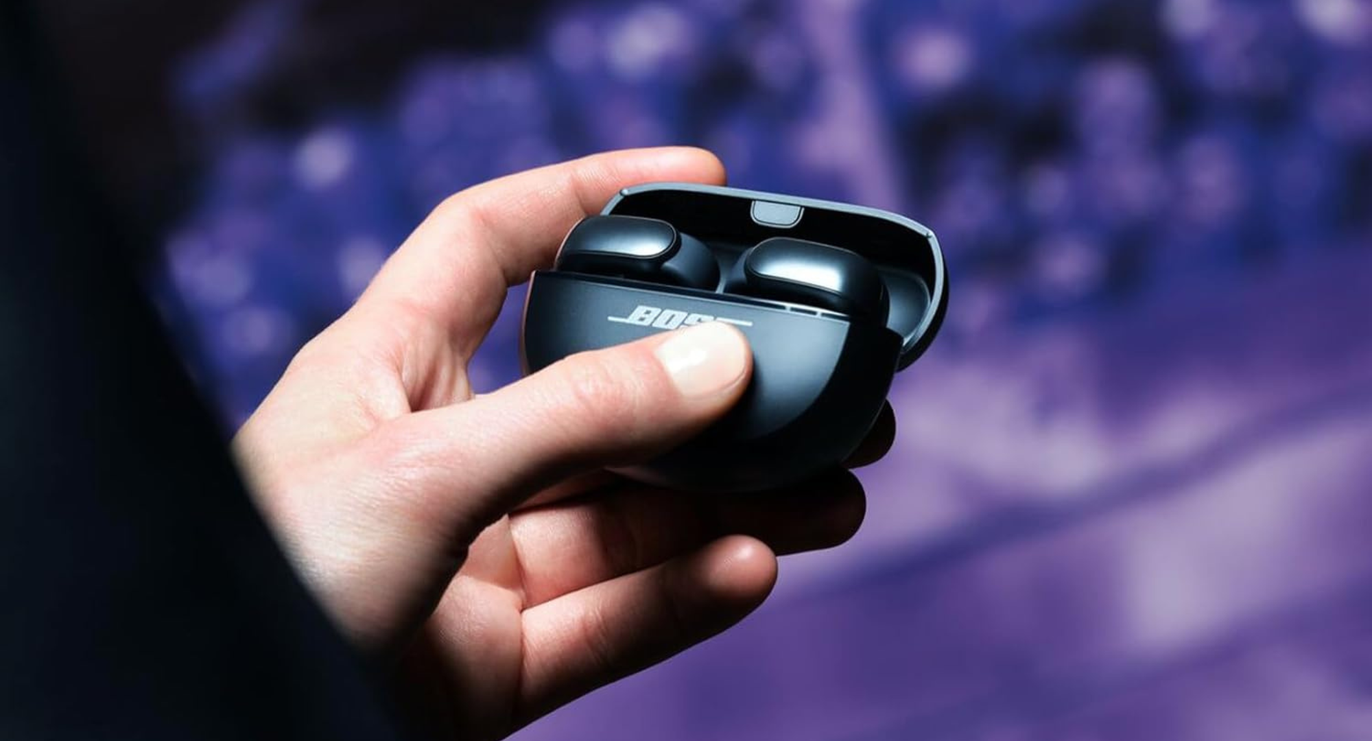 A person's hand holds open a Bose Ultra Open Earbuds charging case, revealing the earbuds inside. The background is blurred with purple and white lights.