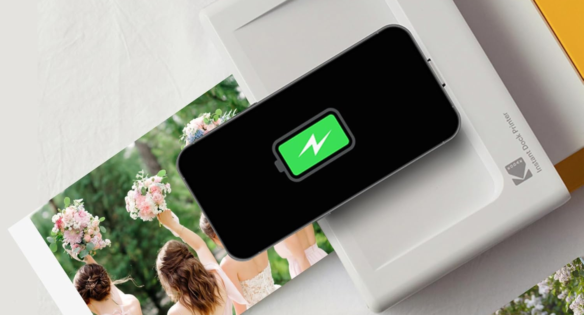 A black smartphone with a green charging battery icon on its screen is docked in a white Kodak Dock Plus 4PASS Instant Photo Printer, with a printed photo showing people holding bouquets visible next to it.