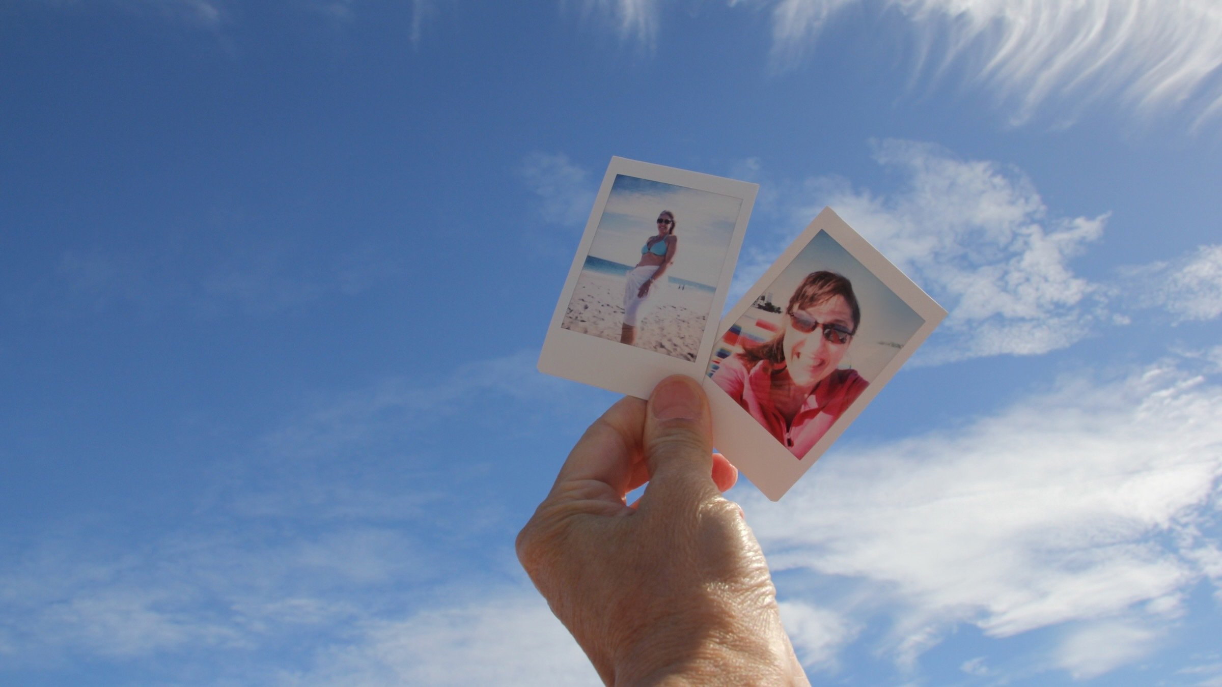 One hand holding up two instant photos of friends against blue sky