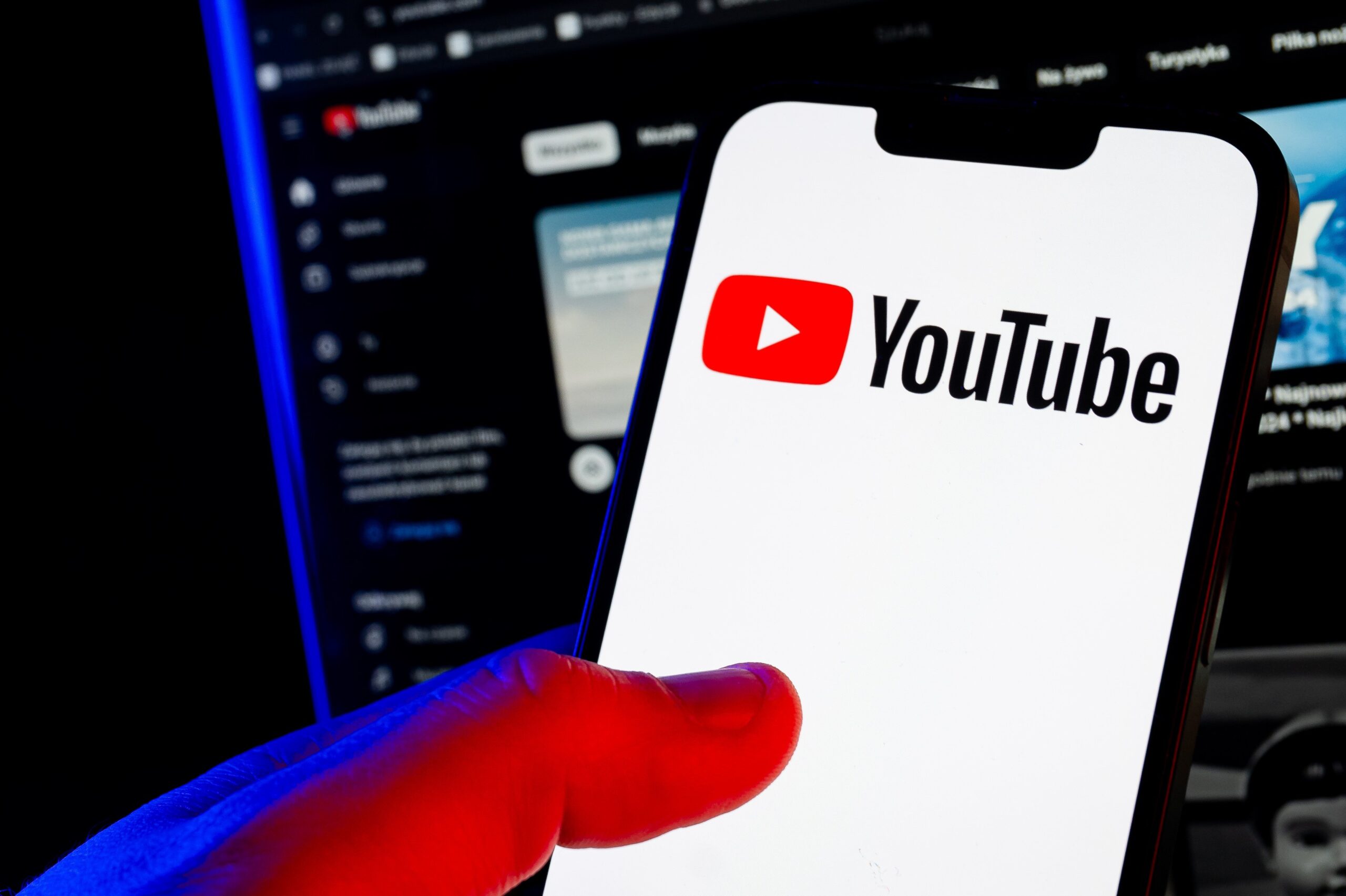 A hand holds a smartphone showing the YouTube logo. A computer screen glows in a darkened room behind it.