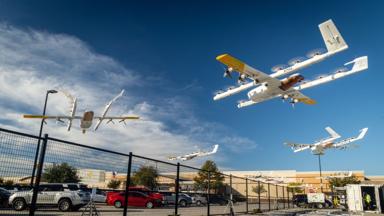Several white and yellow drones fly over a fenced shopping center parking lot.
