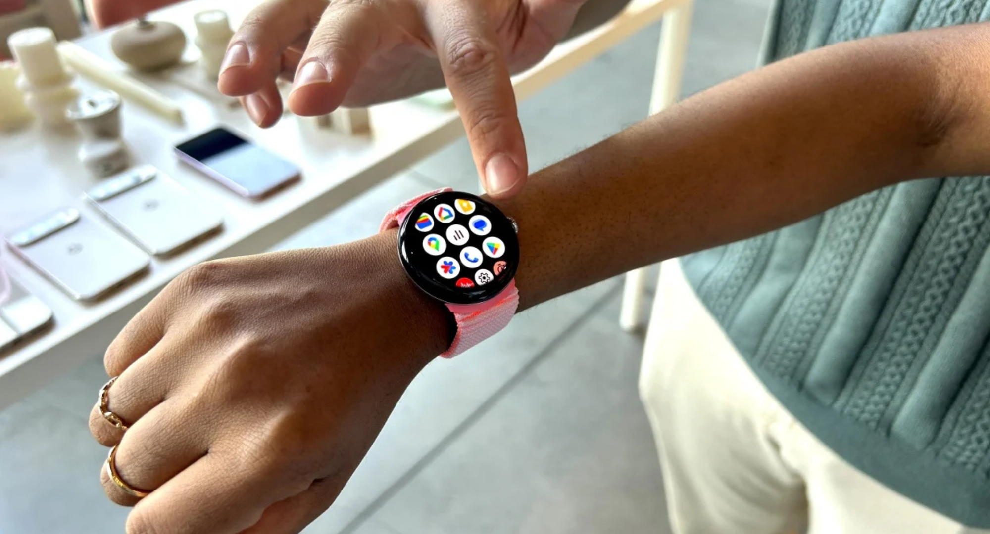 A close-up of a person wearing a black Google Pixel Watch with a pink band, pointing a finger at the screen which displays a grid of app icons.