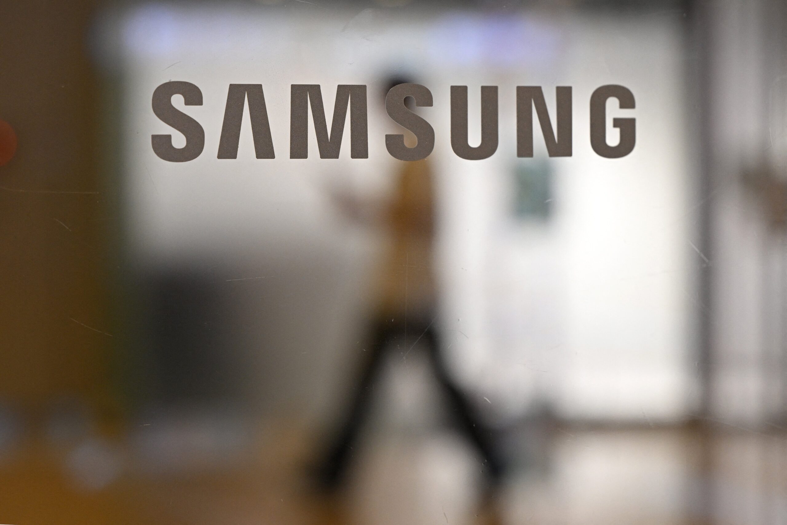 A woman walks past a logo of Samsung Electronics displayed on a glass door at the company's Seocho building in Seoul on July 8, 2025. Samsung Electronics said July 8, it expected its second quarter operating profits to drop 56 percent, blaming US export controls on advanced AI chips to China. 