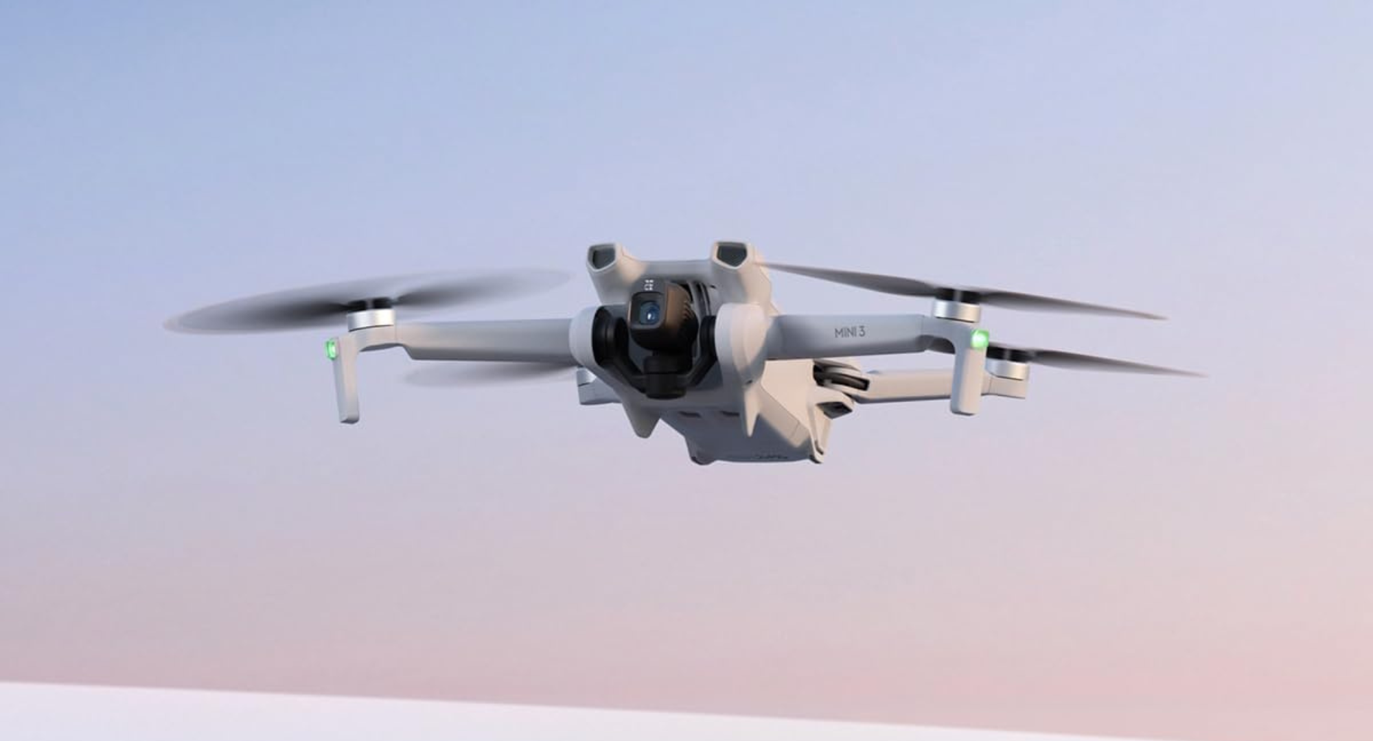 A white DJI Mini 3 drone with its propellers spinning hovers in the air against a soft, light blue and pink sky.