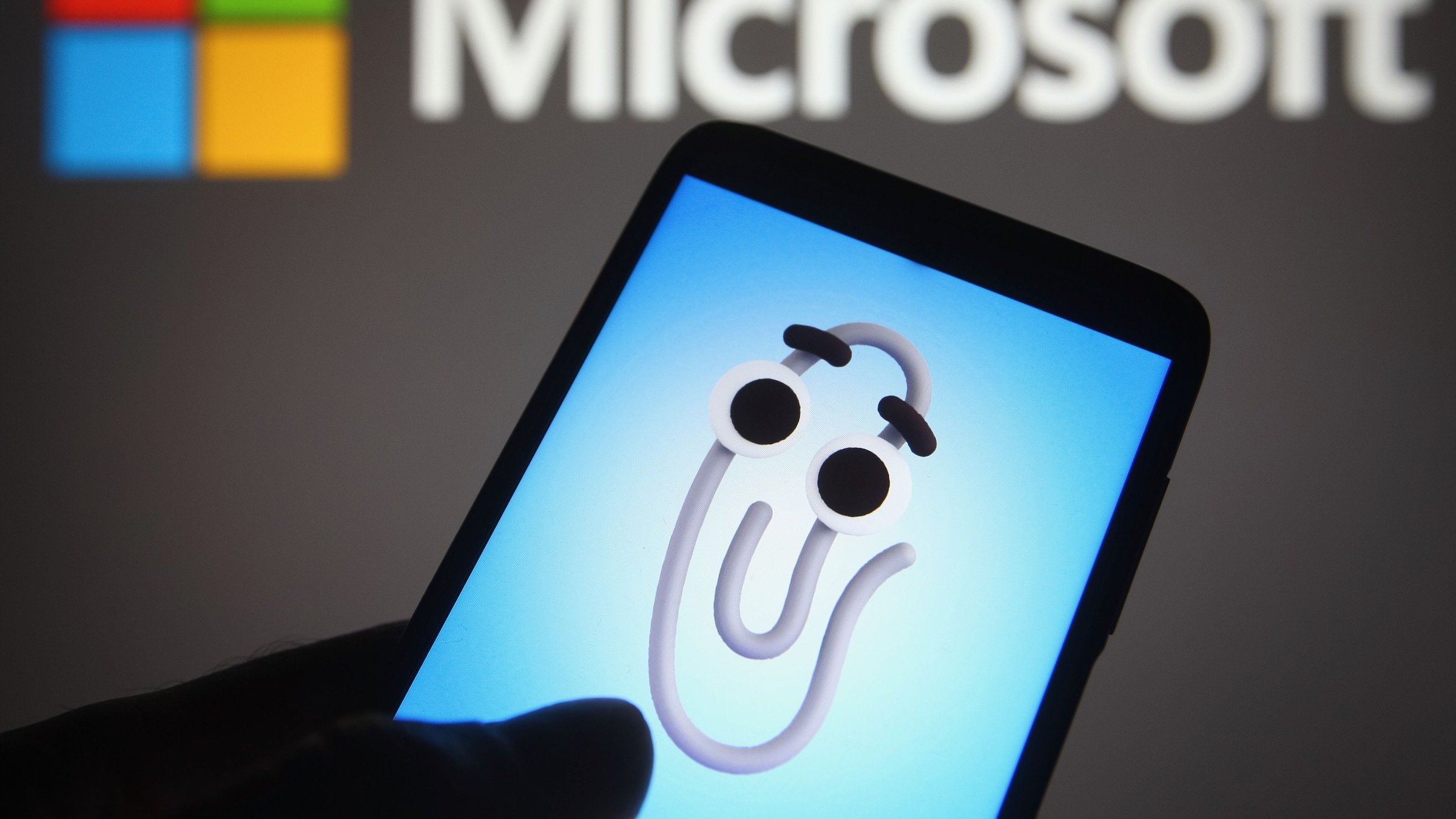 an animated Clippy on a smartphone screen with microsoft logo in background