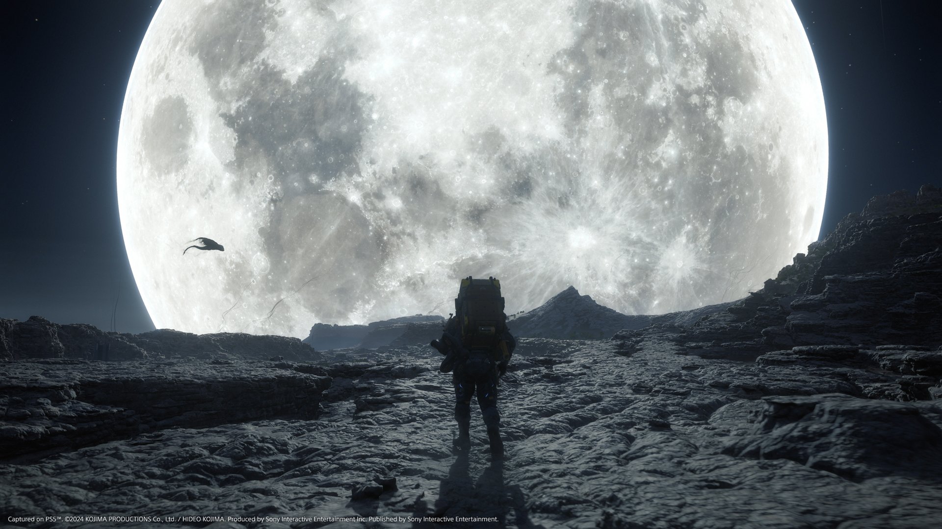 A screenshot from the video game 'Death Stranding' showing a person standing on a barren landscape in front of a giant, rising full moon